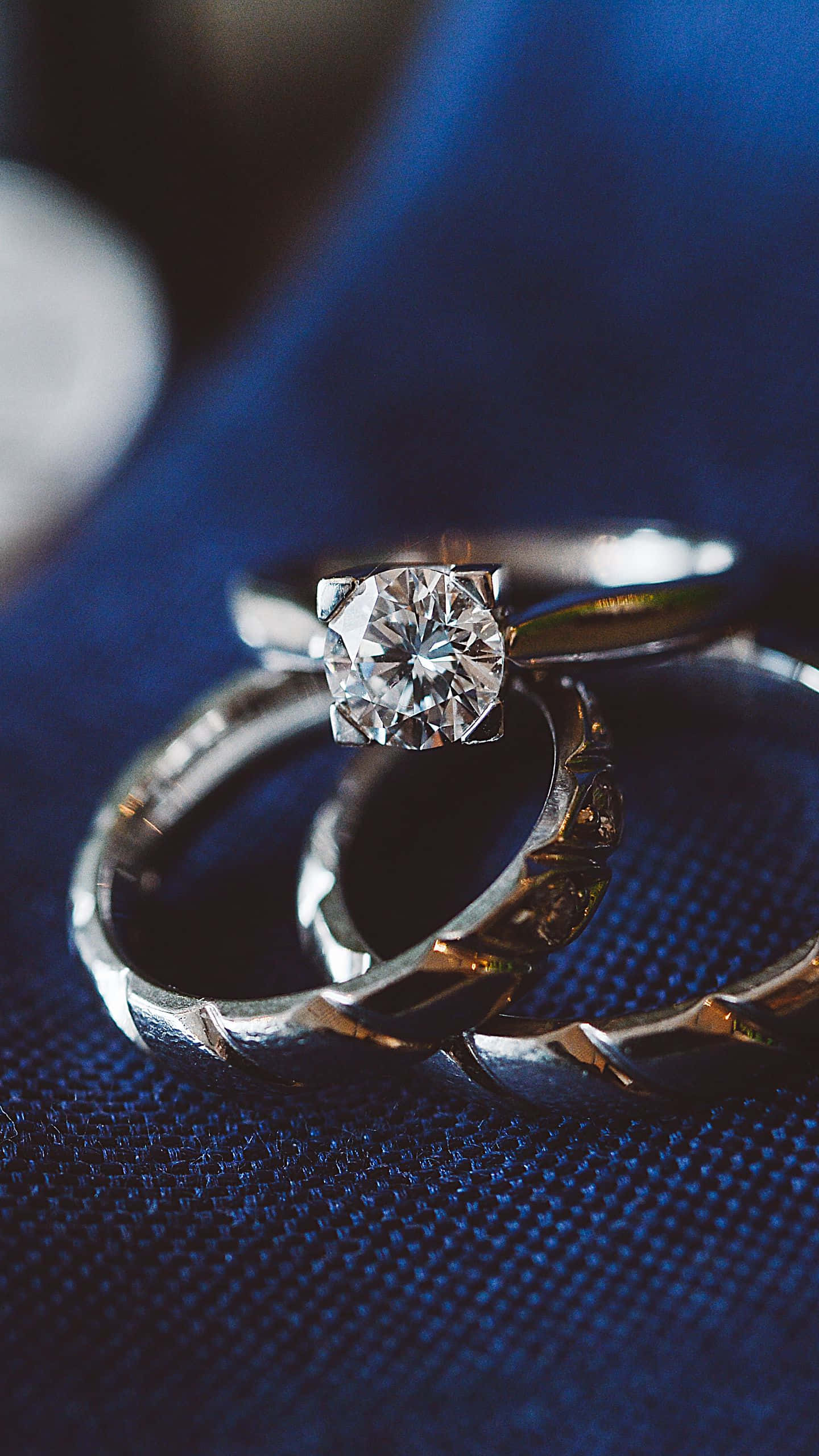 Wedding Ring 桌布 壁紙 — 兩枚結婚戒指，其中一枚鑲有大鑽石，放在有紋理的藍色布料上，呈現柔焦效果。