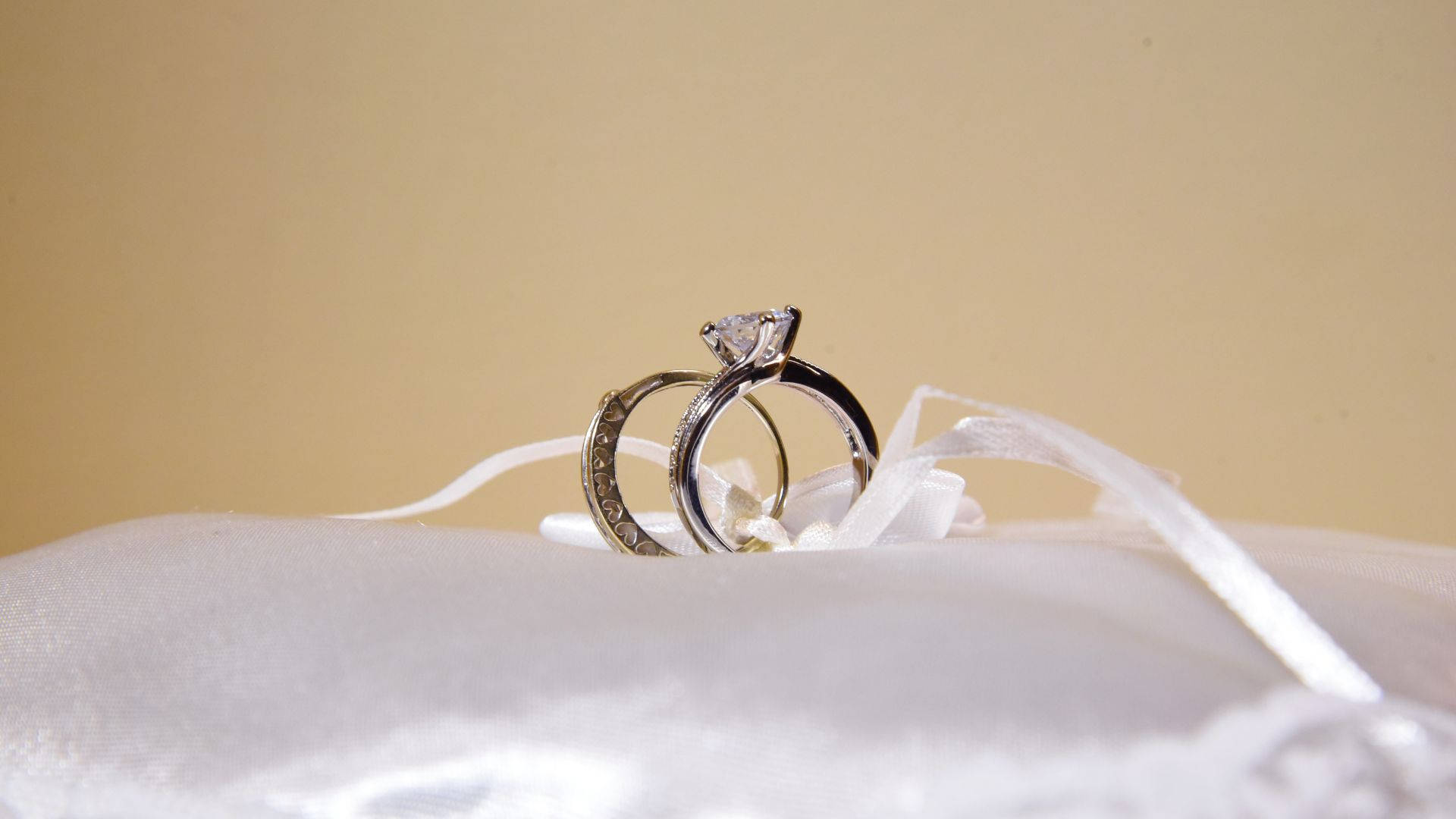 高清 桌布 壁紙 — 兩枚結婚戒指，其中一枚鑲有大鑽石，放在白色緞面枕頭上，背景為柔和米色。