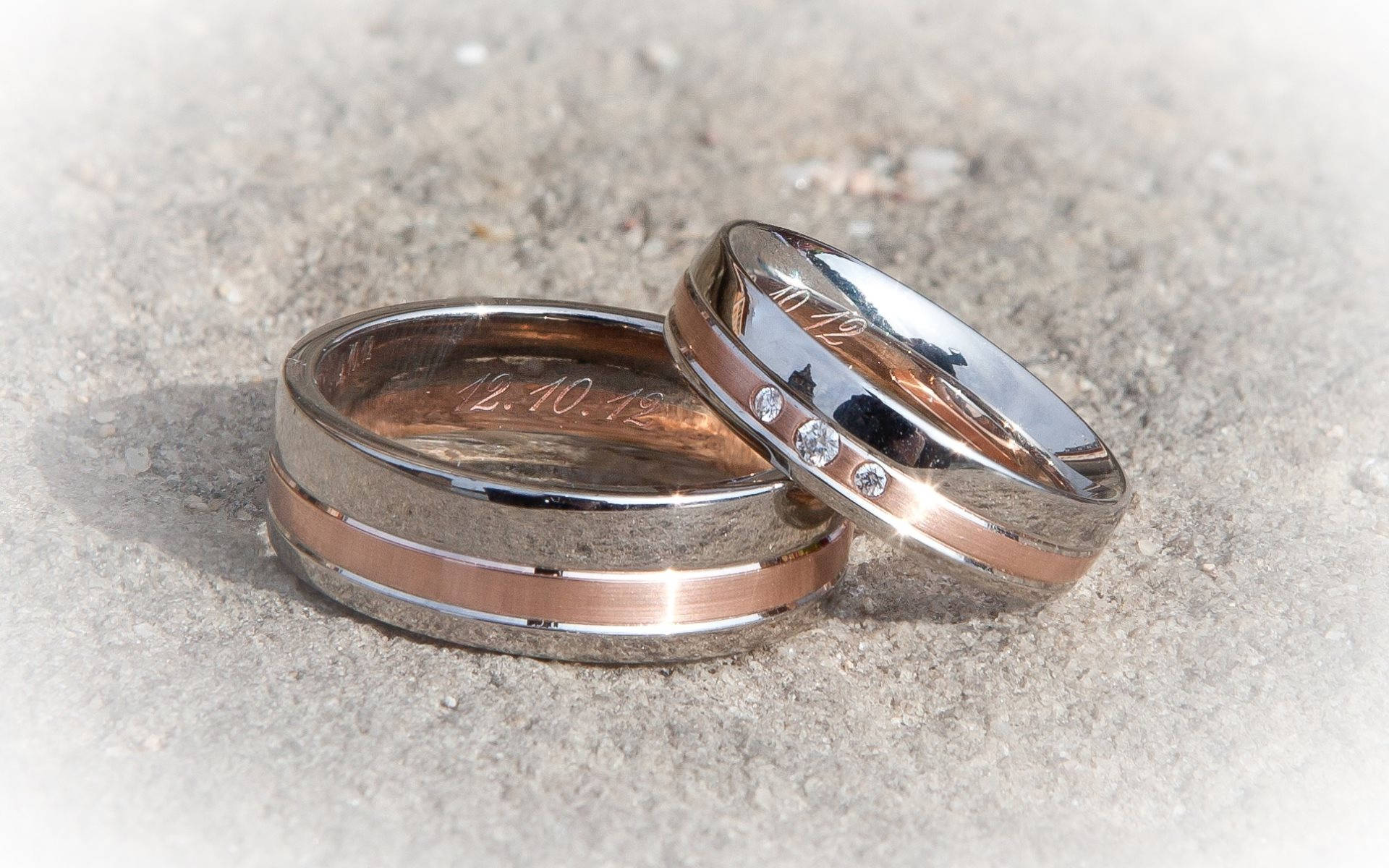 Wedding Rings 桌布 壁紙 — 兩枚結婚戒指，其中一枚鑲有鑽石並刻有日期，放在紋理混凝土上，在明亮的陽光下。
