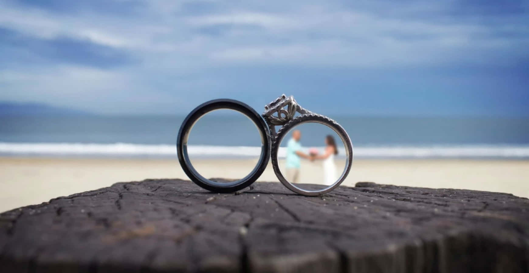 結婚戒指 桌布 壁紙 — 兩枚結婚戒指放在一根風化的木頭上，背景是沙灘上模糊的情侶牽手畫面。