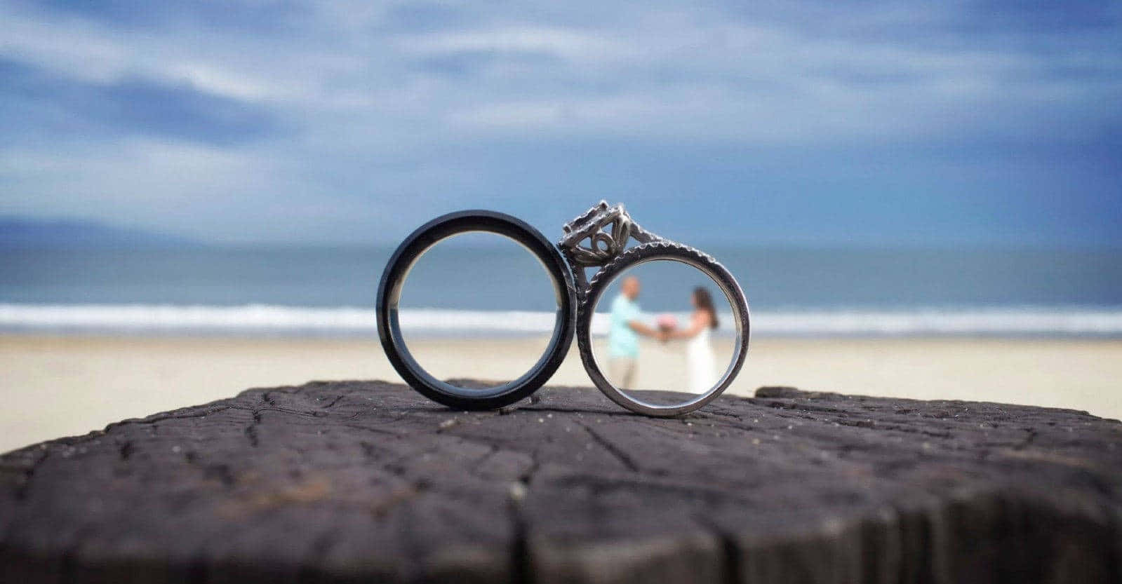Wedding Rings 桌布 壁紙 — 兩枚結婚戒指放在一條經風吹日曬的木頭上，背景是一對模糊的情侶在海灘上牽手。