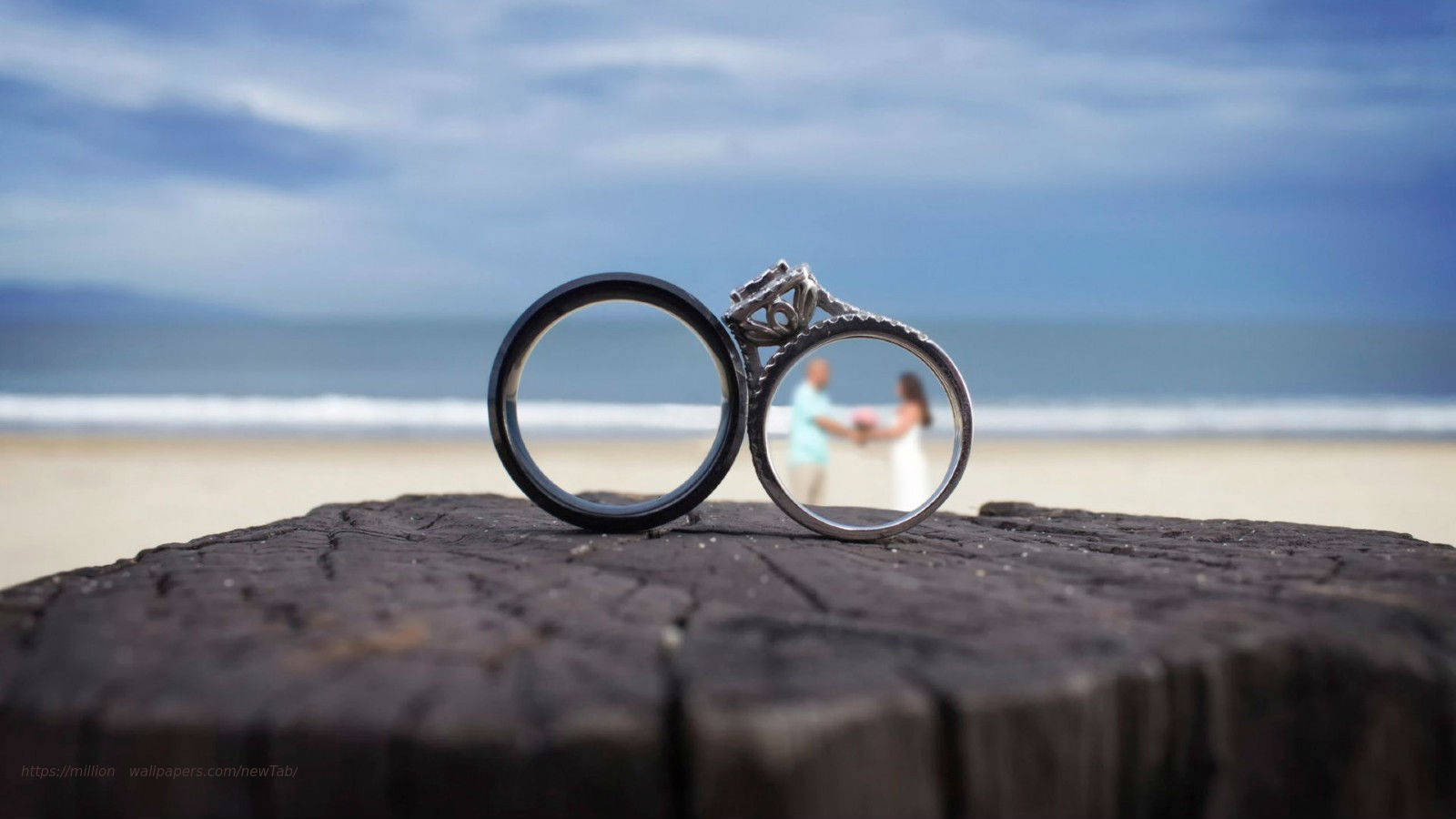 Wedding Rings 桌布 壁紙 — 兩枚結婚戒指放在一條風化的木頭上，背景是一對模糊的情侶在海灘上牽手。