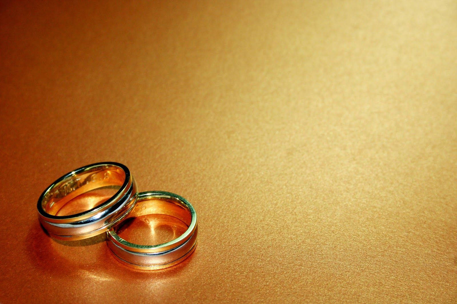 Wedding Rings 桌布 壁紙 — 兩枚金婚戒一起放在溫暖有紋理的金色表面上，光線柔和。