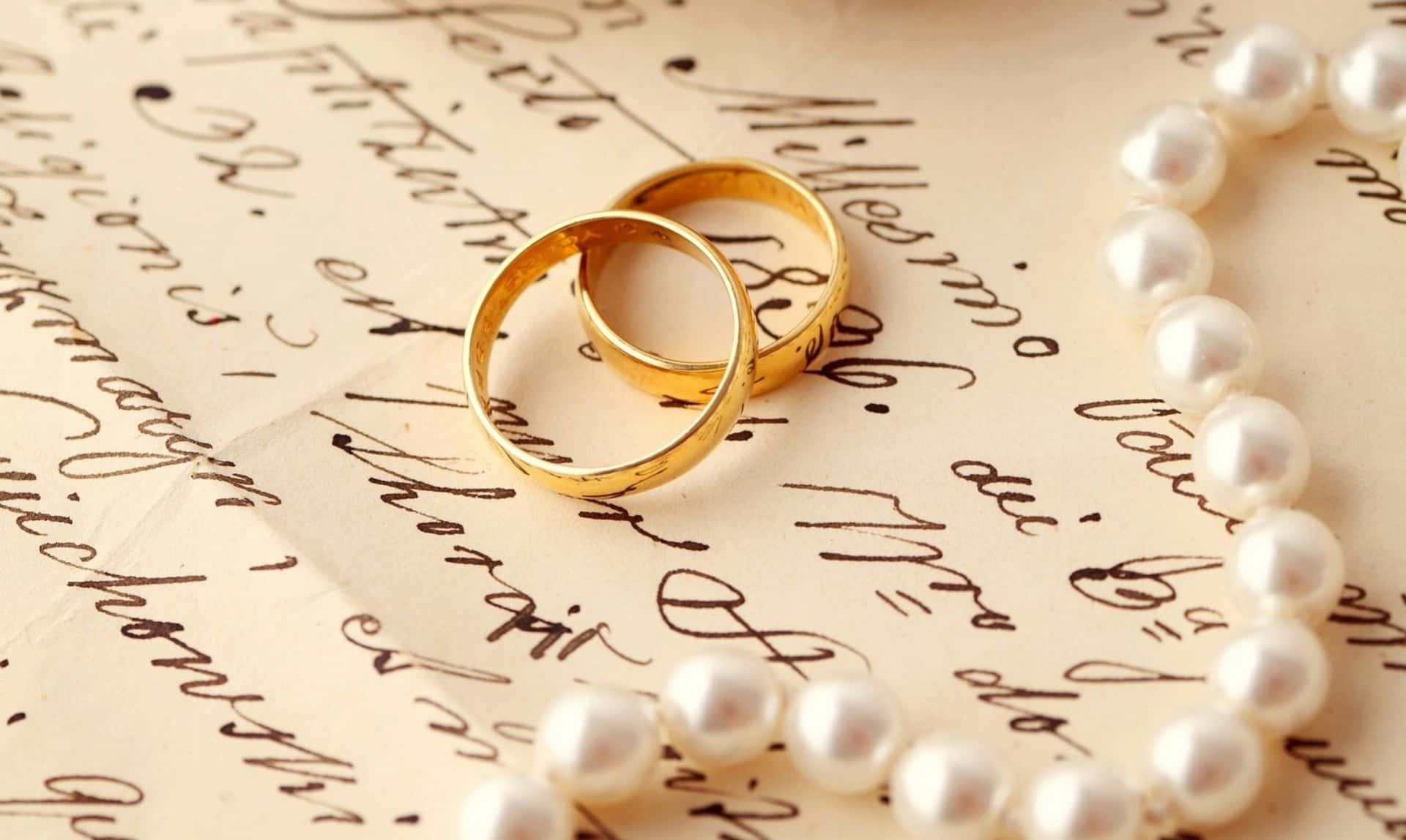 結婚戒指 桌布 壁紙 — 兩枚金婚戒和一條珍珠項鍊優雅地放置在復古手寫字紙上。