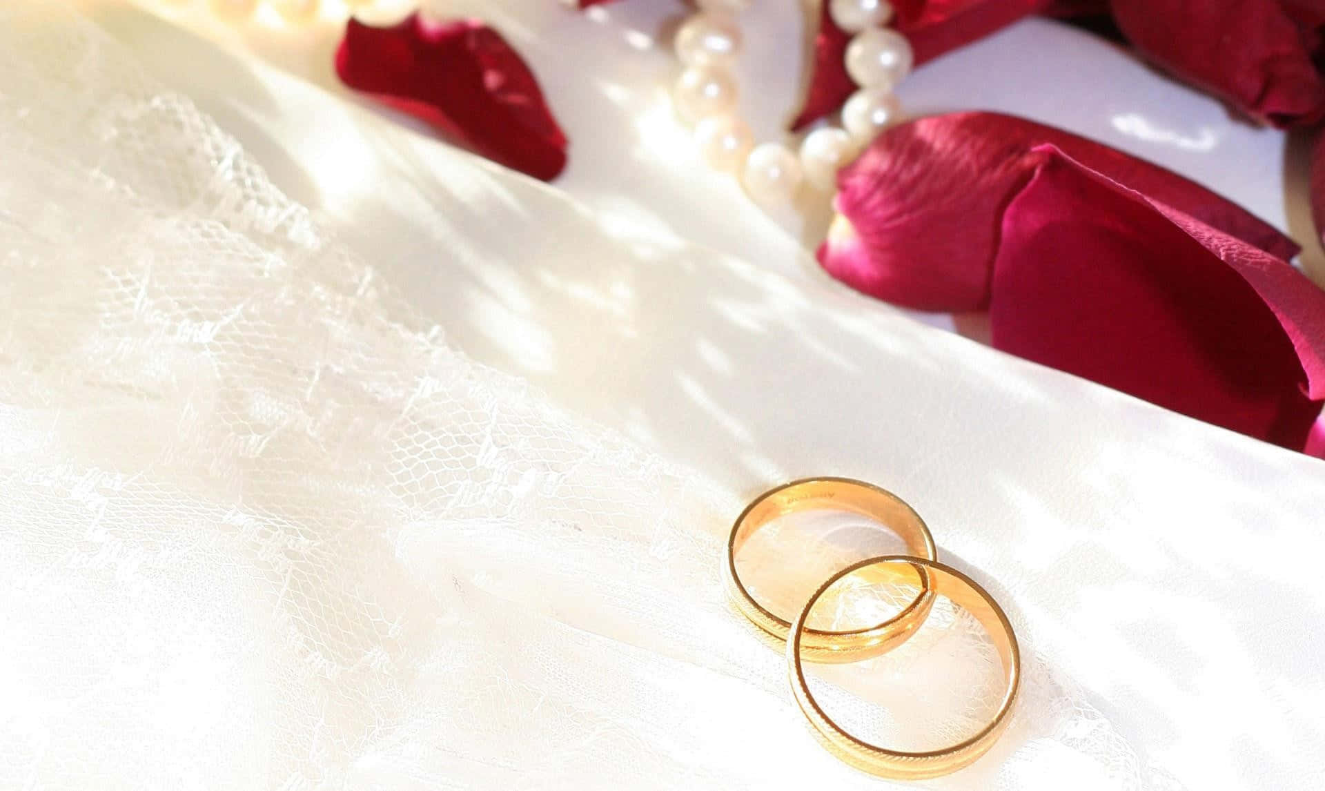 結婚戒指 桌布 壁紙 — 兩枚金婚戒放在白色蕾絲上，旁邊有紅玫瑰花瓣和珍珠項鍊，在柔和陽光下。