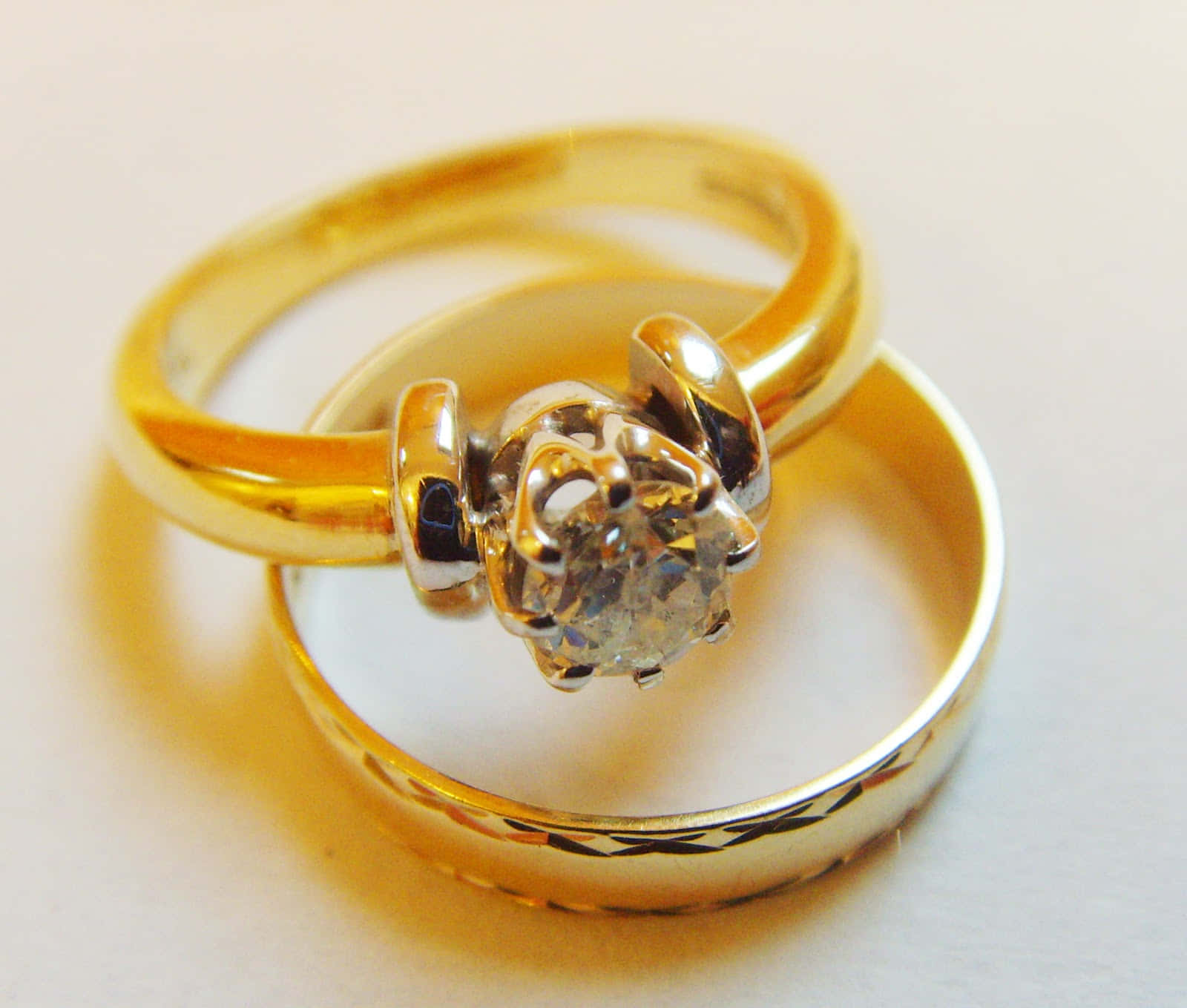 Wedding Rings 桌布 壁紙 — 兩枚金結婚戒指，其中一枚鑲有單鑽，交纏在柔軟的白色表面上。