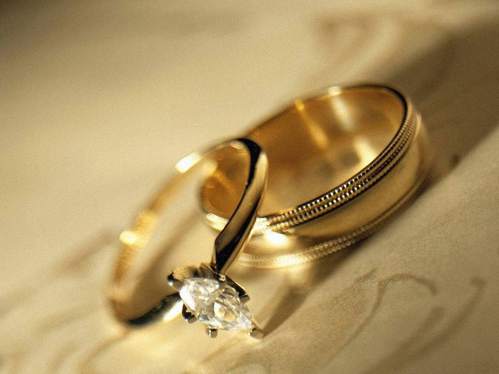 Wedding Rings 桌布 壁紙 — 兩枚金結婚戒指，其中一枚鑲有馬眼形鑽石，放在柔軟的奶油色布料上，沐浴在溫暖的金色光線中。