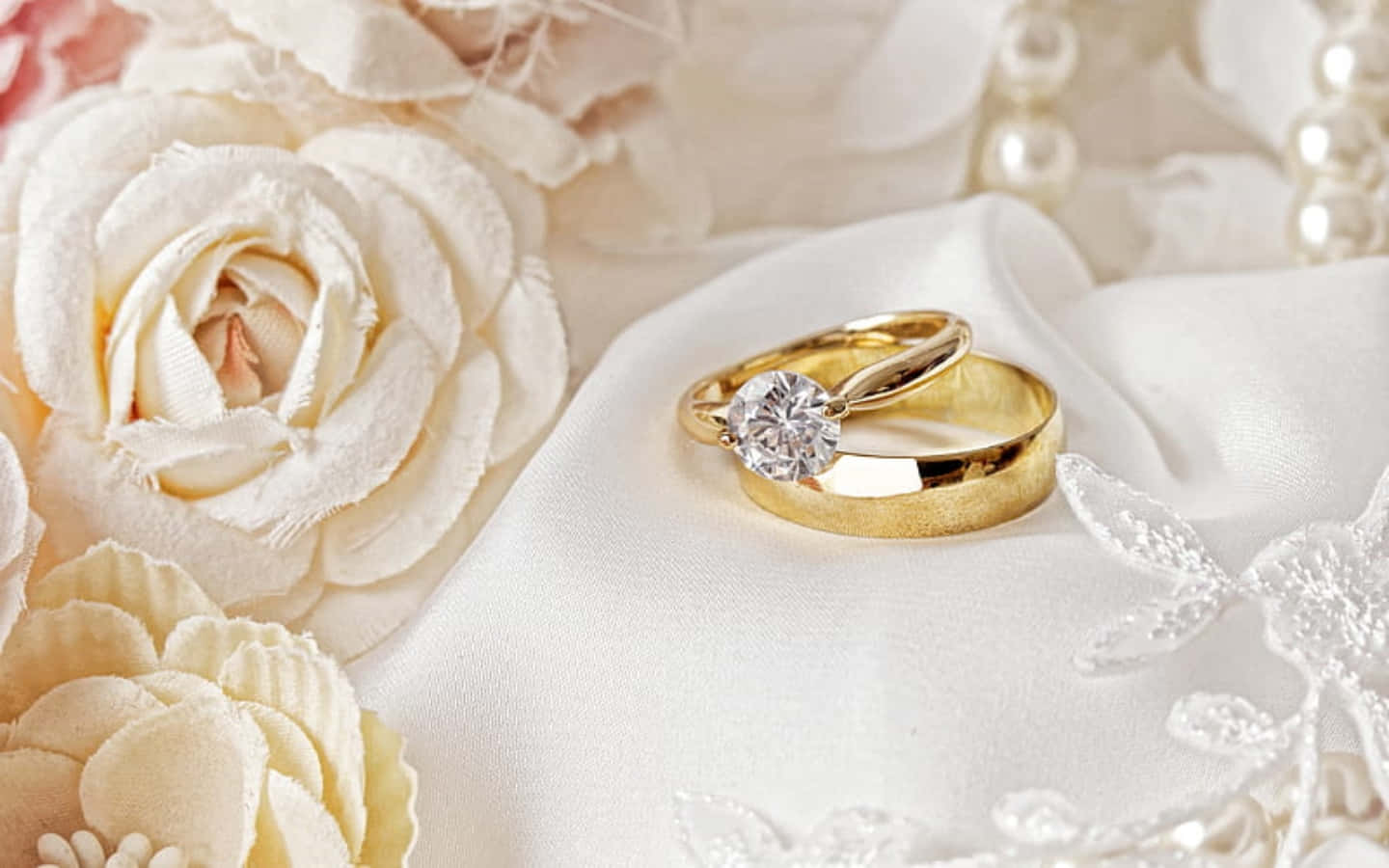 結婚戒指 桌布 壁紙 — 兩枚金結婚戒指，鑲有鑽石，放在白色絲綢上，旁邊有奶油色玫瑰和珍珠。
