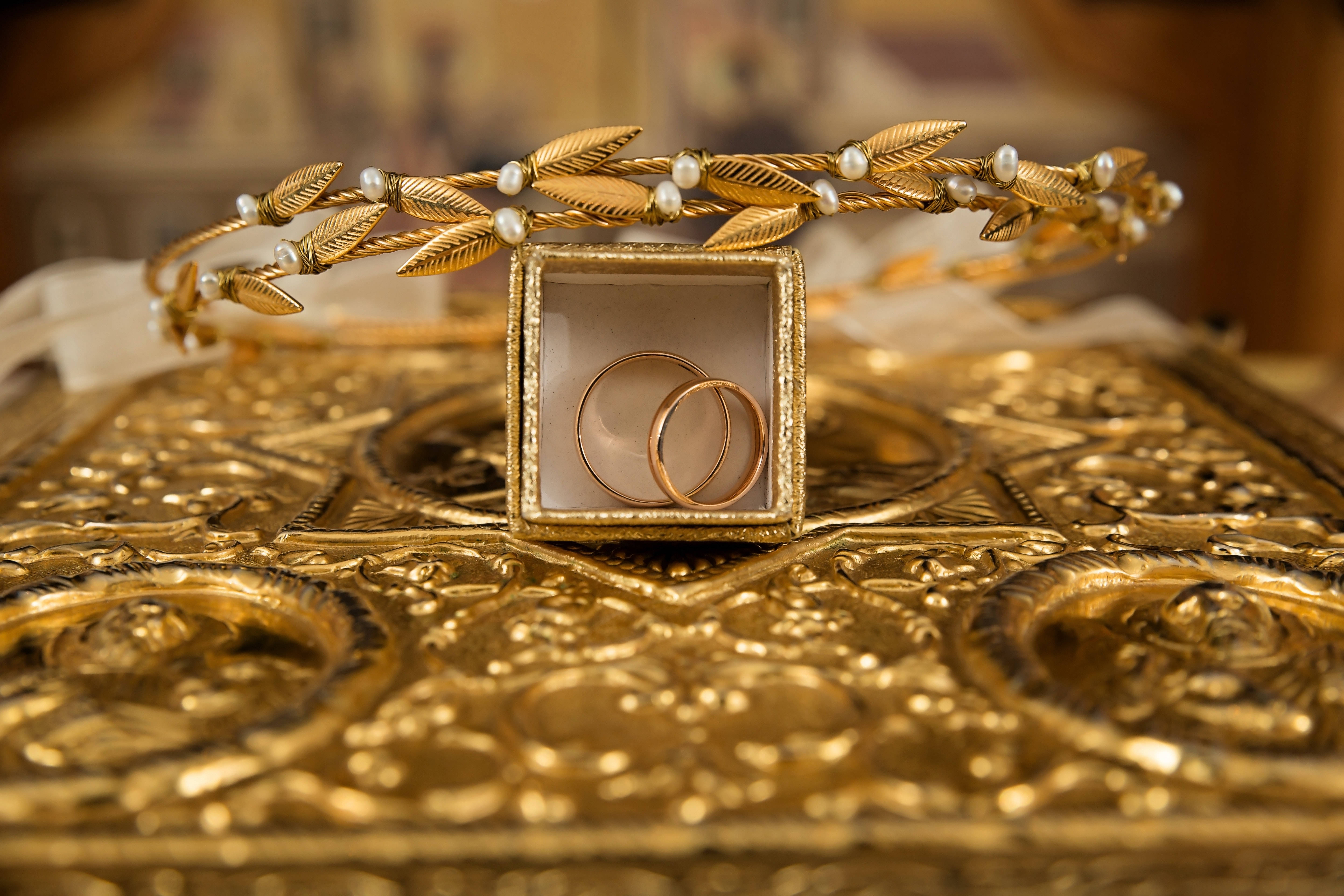 Gold Jewellery 桌布 壁紙 — 兩枚金結婚戒指放在一個盒子裡，置於華麗的金色表面上，旁邊有一個樹葉和珍珠皇冠。