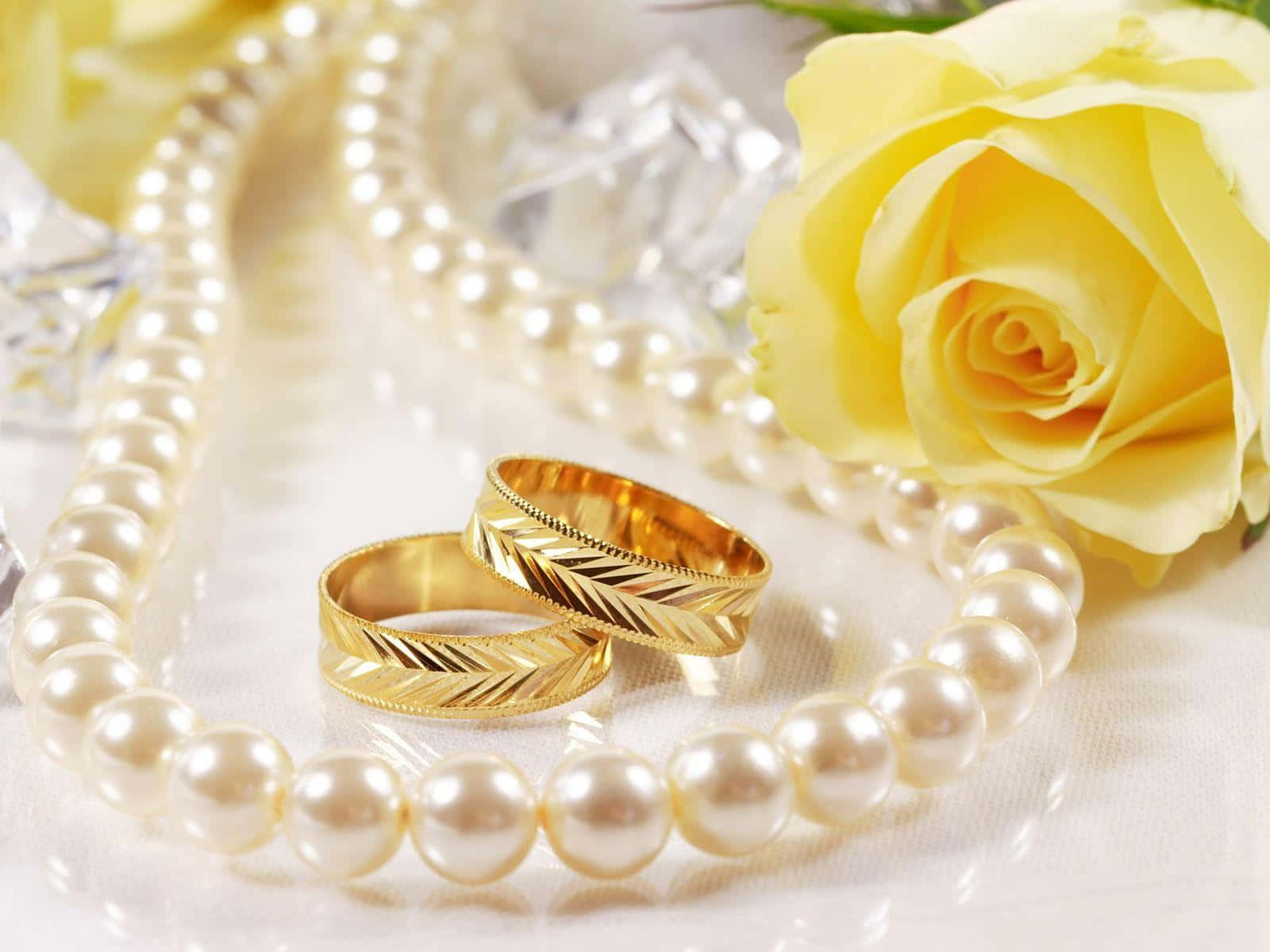 Ring 桌布 壁紙 — 兩枚金結婚戒指放在珍珠項鍊上，旁邊有黃玫瑰和水晶，散發優雅與浪漫。
