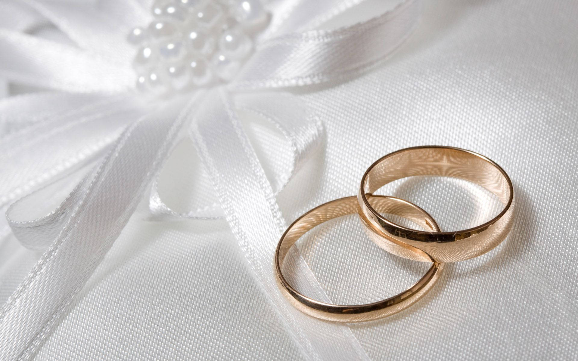 Wedding Rings 桌布 壁紙 — 兩枚金結婚戒指放在白色緞面表面，旁邊有一個珍珠裝飾的蝴蝶結。