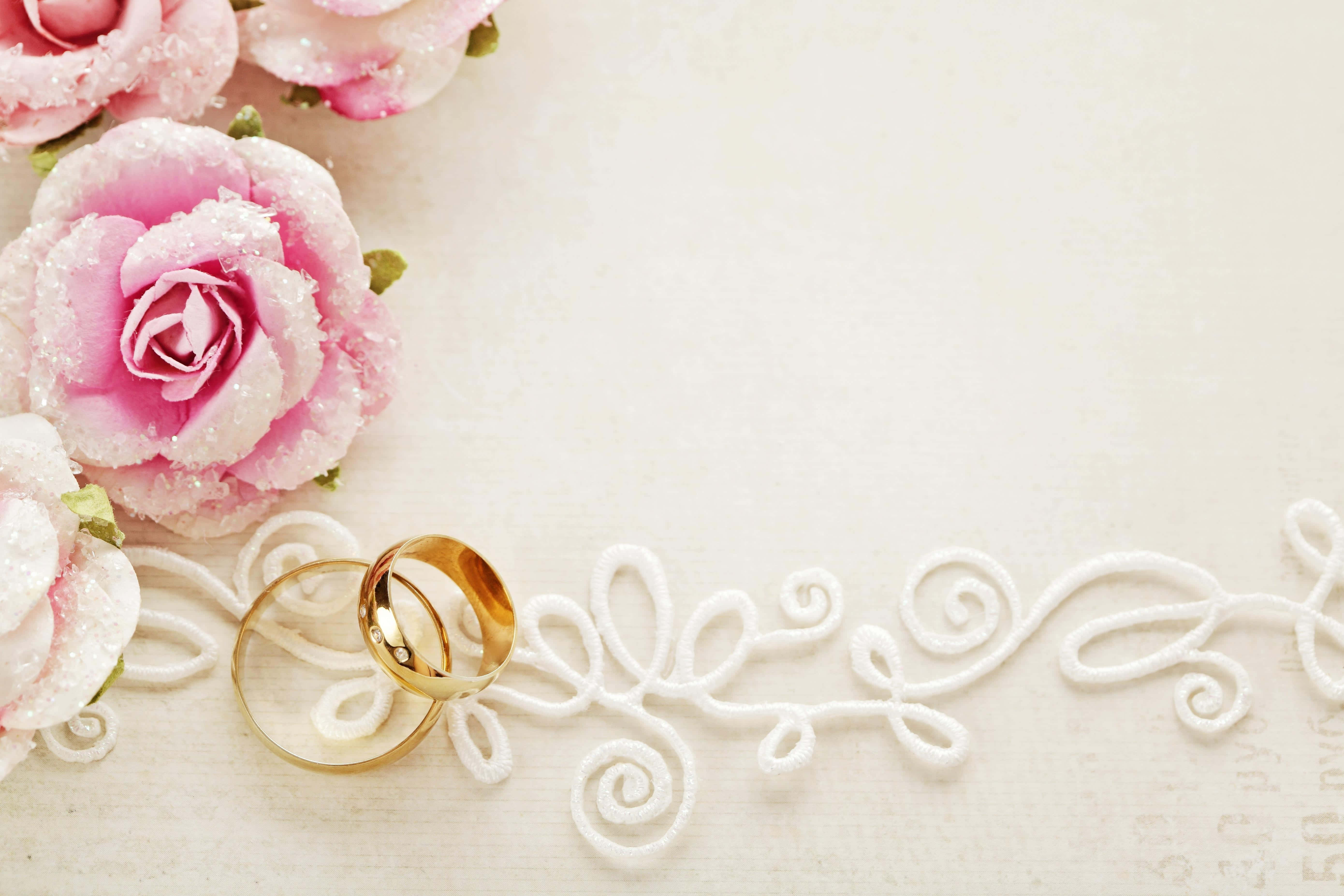 Wedding Rings 桌布 壁紙 — 兩枚金結婚戒指放在白色蕾絲上，旁邊是撒有亮粉的粉紅玫瑰，背景是柔和的奶油色。