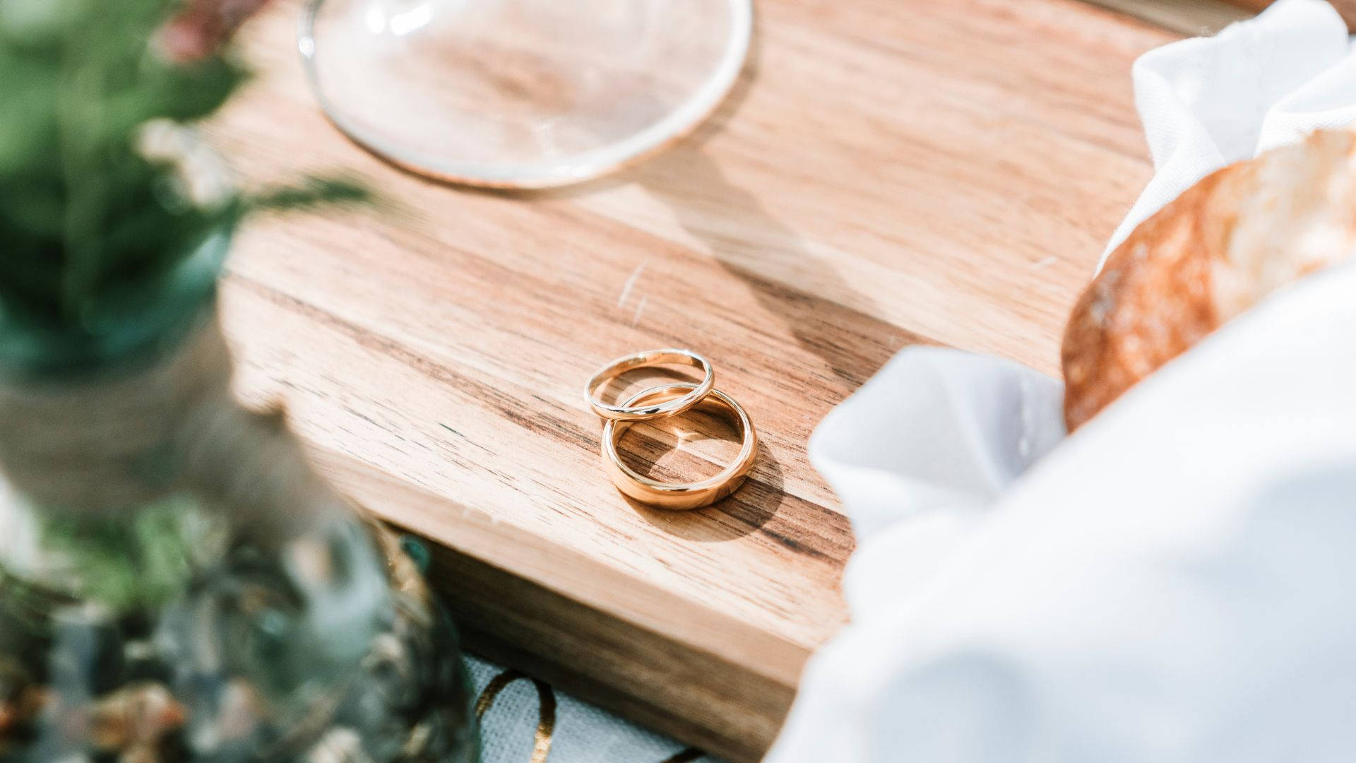 Wedding Rings 桌布 壁紙 — 兩枚金結婚戒指放在陽光照射的木托盤上，旁邊有玻璃杯和白色布。