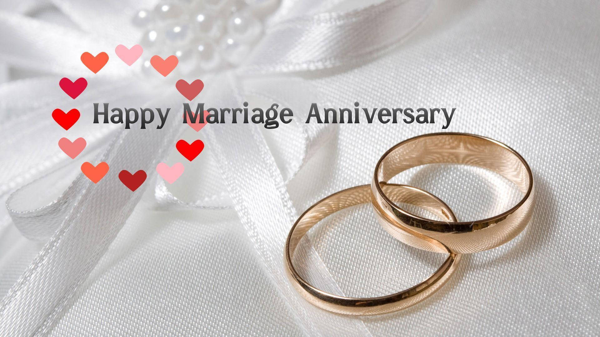 Happy Anniversary 桌布 壁紙 — 兩枚金色結婚戒指交織在白色緞帶上，帶有心形圖案和週年紀念文字。