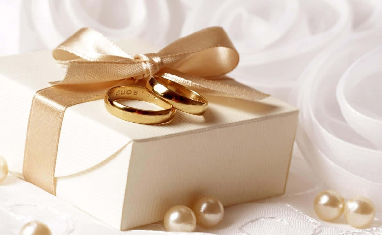 Wedding Rings 桌布 壁紙 — 兩枚金色結婚戒指放在一個用金色絲帶綁著的奶油色禮物盒上，周圍環繞著珍珠和白色布料。