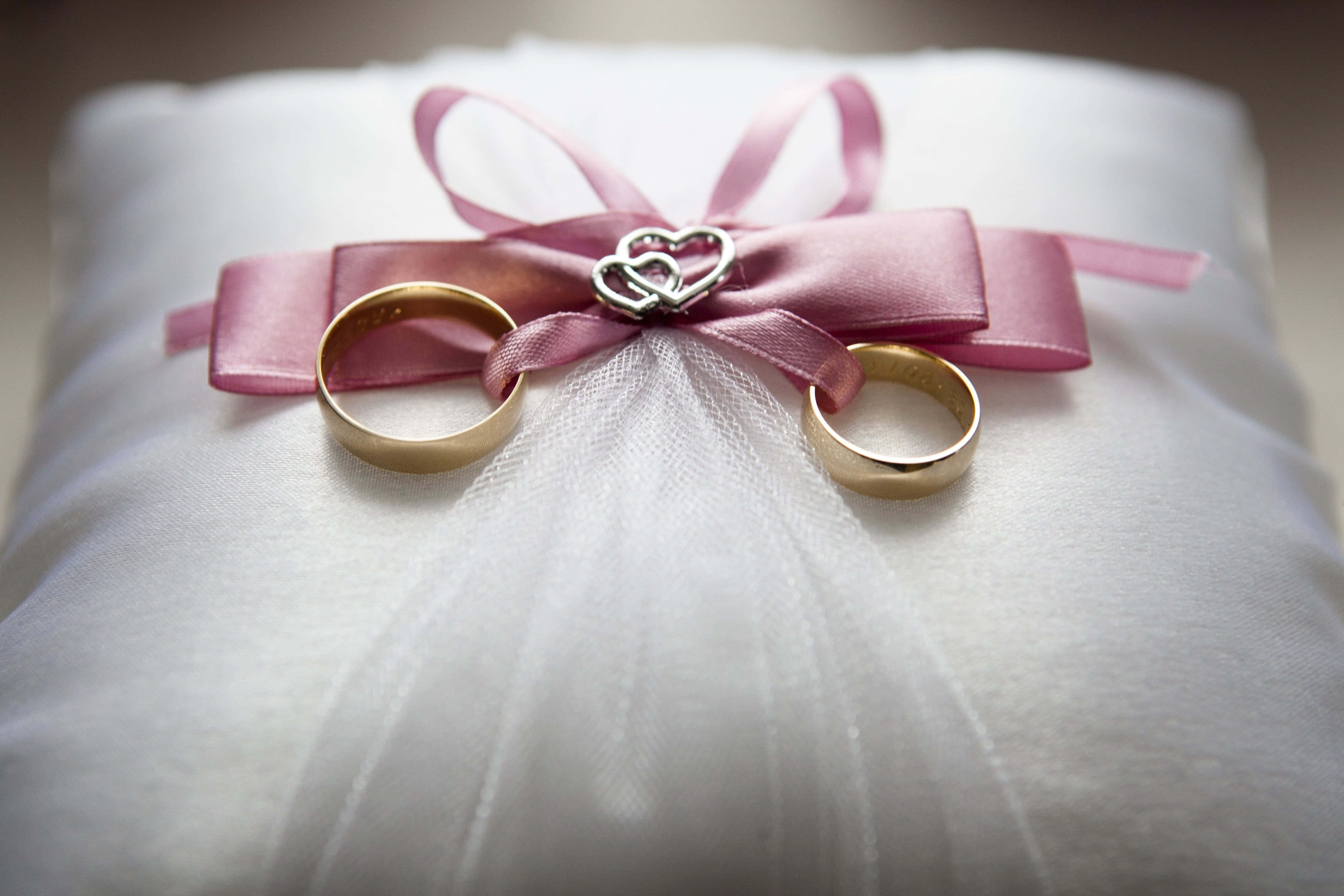 Wedding Rings 桌布 壁紙 — 兩枚金色結婚戒指放在一個白色薄紗枕頭上，枕頭繫有粉紅色緞帶蝴蝶結和心形吊飾。