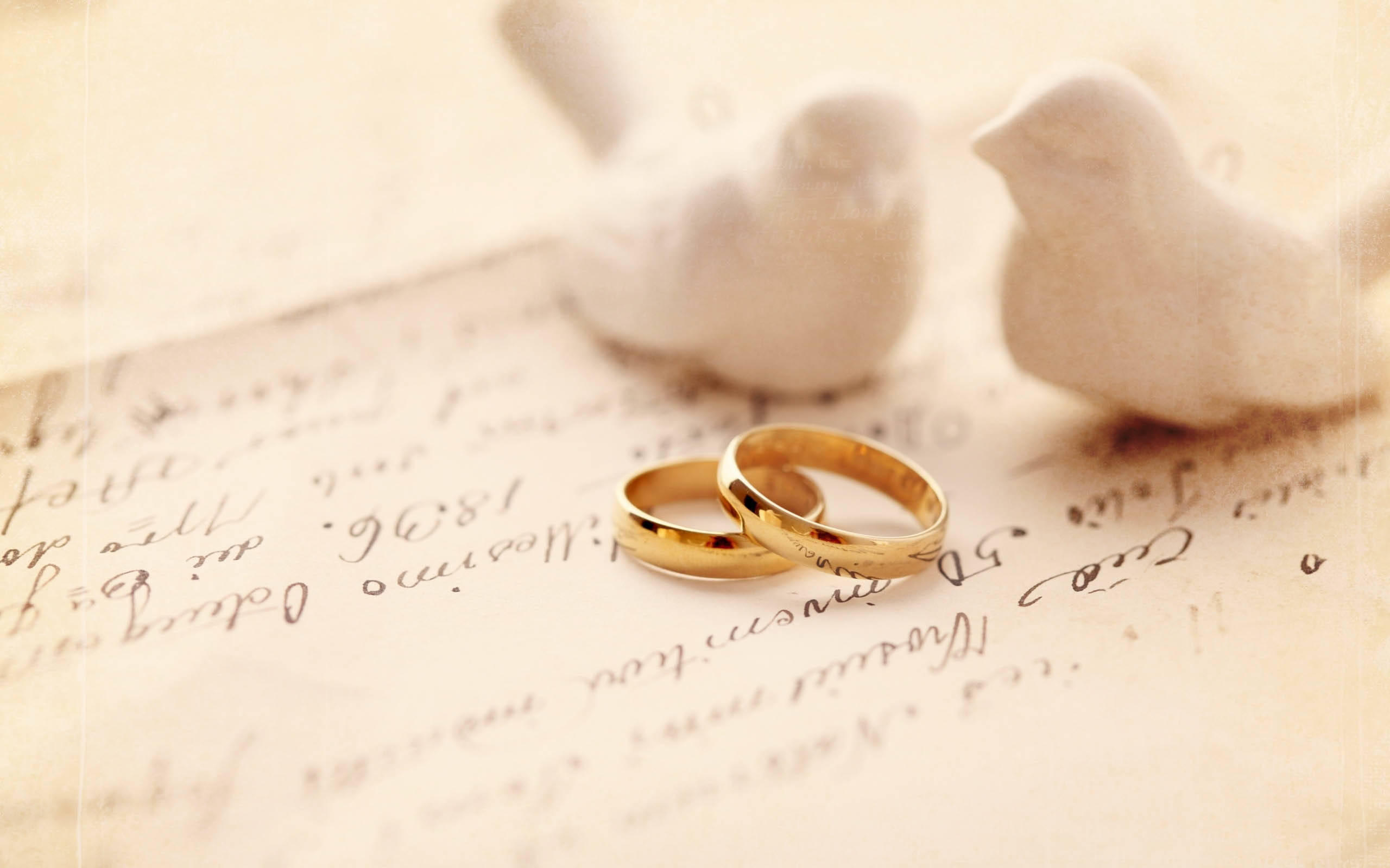 birds 桌布 壁紙 — 兩枚金色結婚戒指放在復古手寫紙上，旁邊有白色陶瓷鳥，在柔和溫暖的光線中。