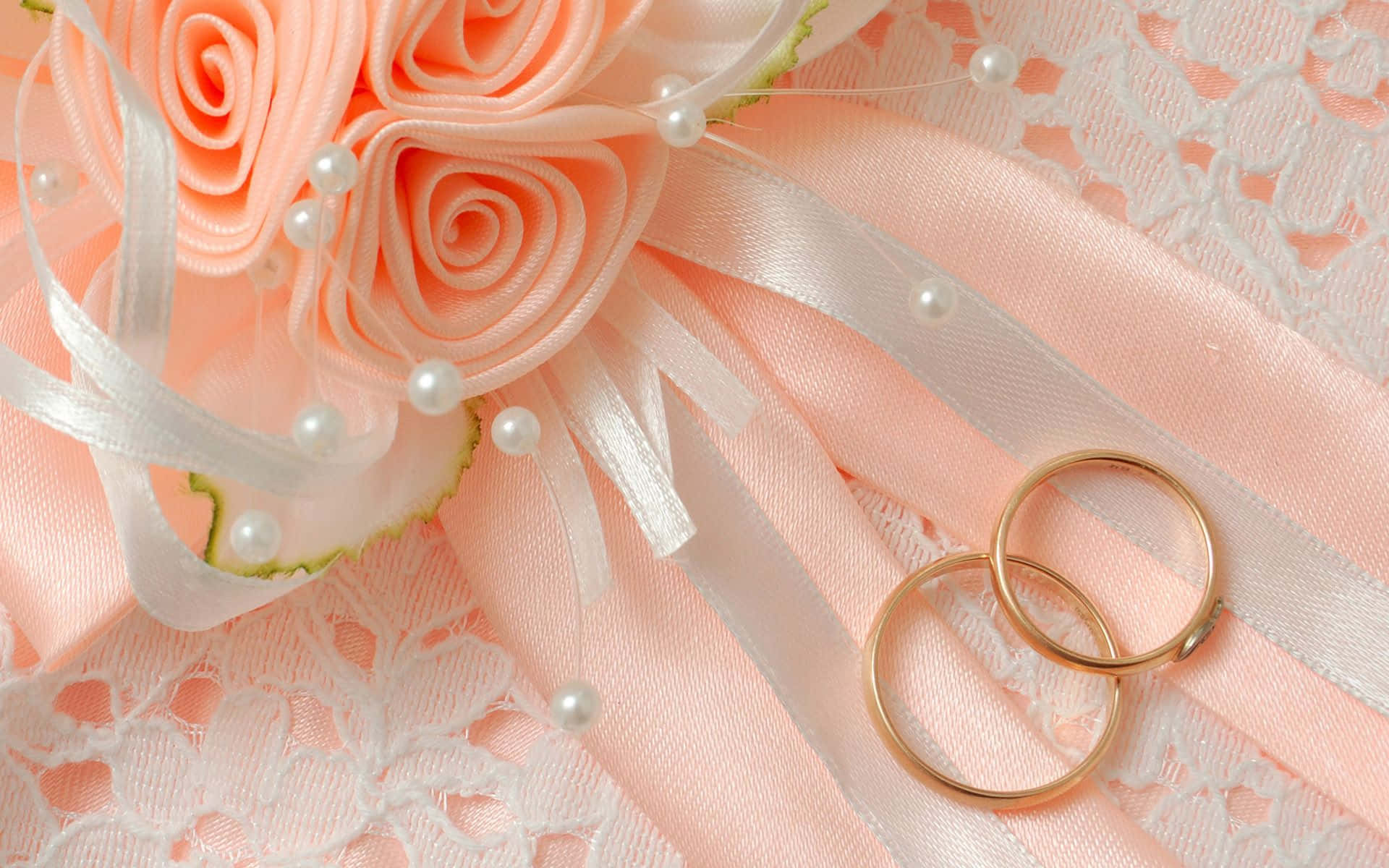 Wedding Flowers 桌布 壁紙 — 兩枚金色結婚戒指放在桃色緞面玫瑰和白色蕾絲旁邊，背景是柔和的粉彩。