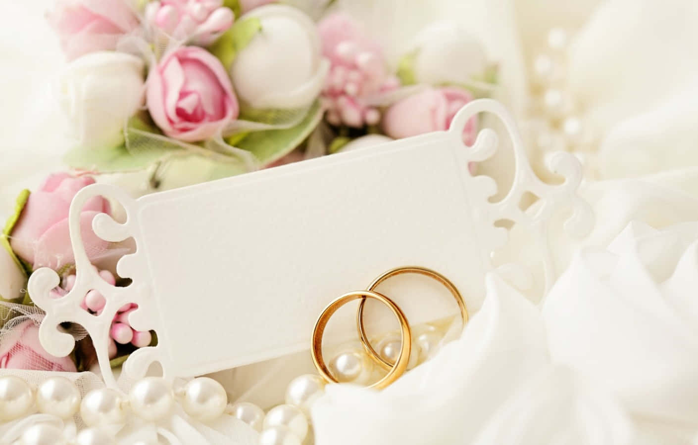結婚戒指 桌布 壁紙 — 兩枚金色結婚戒指放在珍珠上，旁邊有一張白色卡片和柔和的粉白色花朵，構成浪漫的新娘佈置。