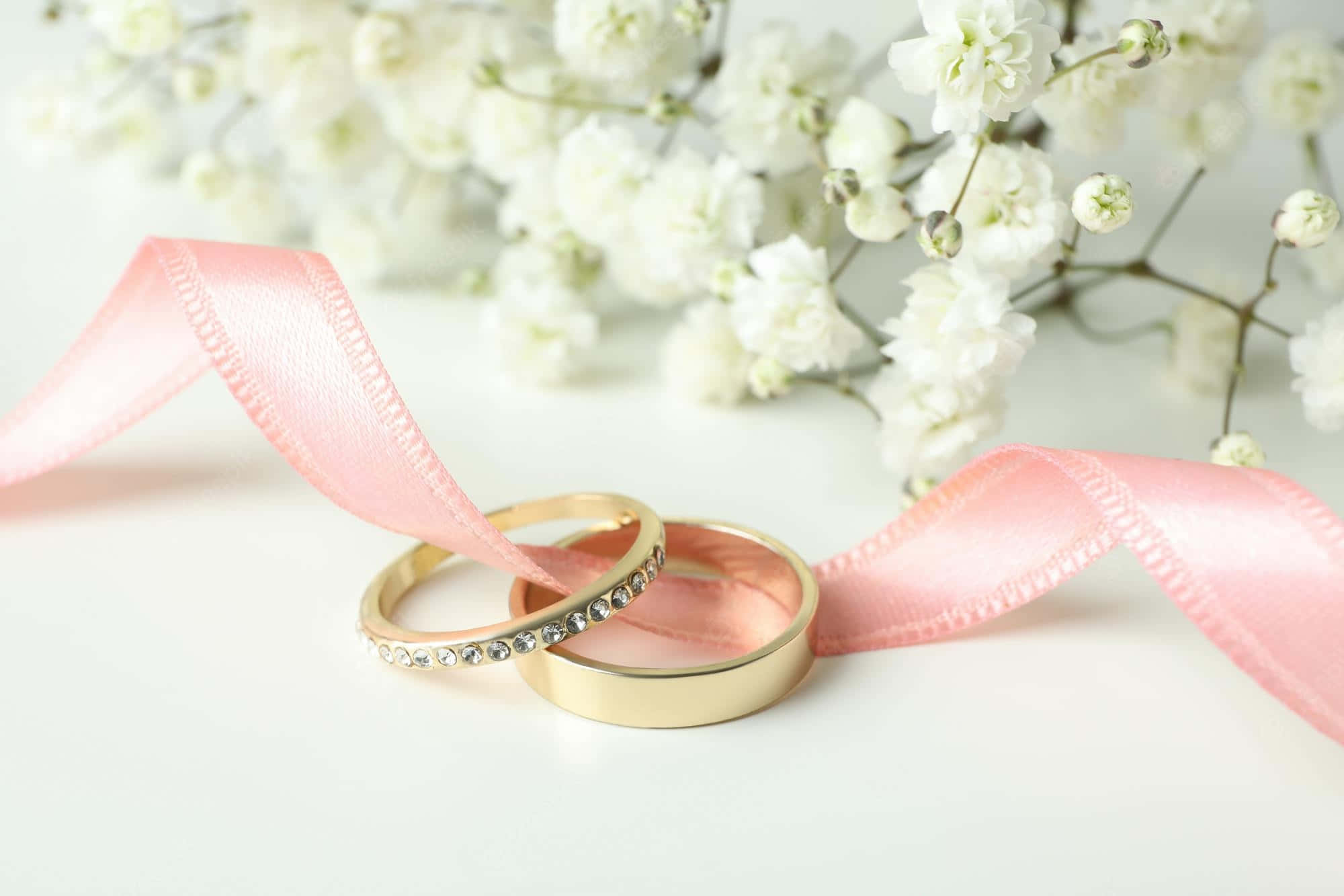 Wedding 桌布 壁紙 — 兩枚金色結婚戒指纏繞著柔軟的粉紅色絲帶，背景是精緻的白色滿天星花朵。
