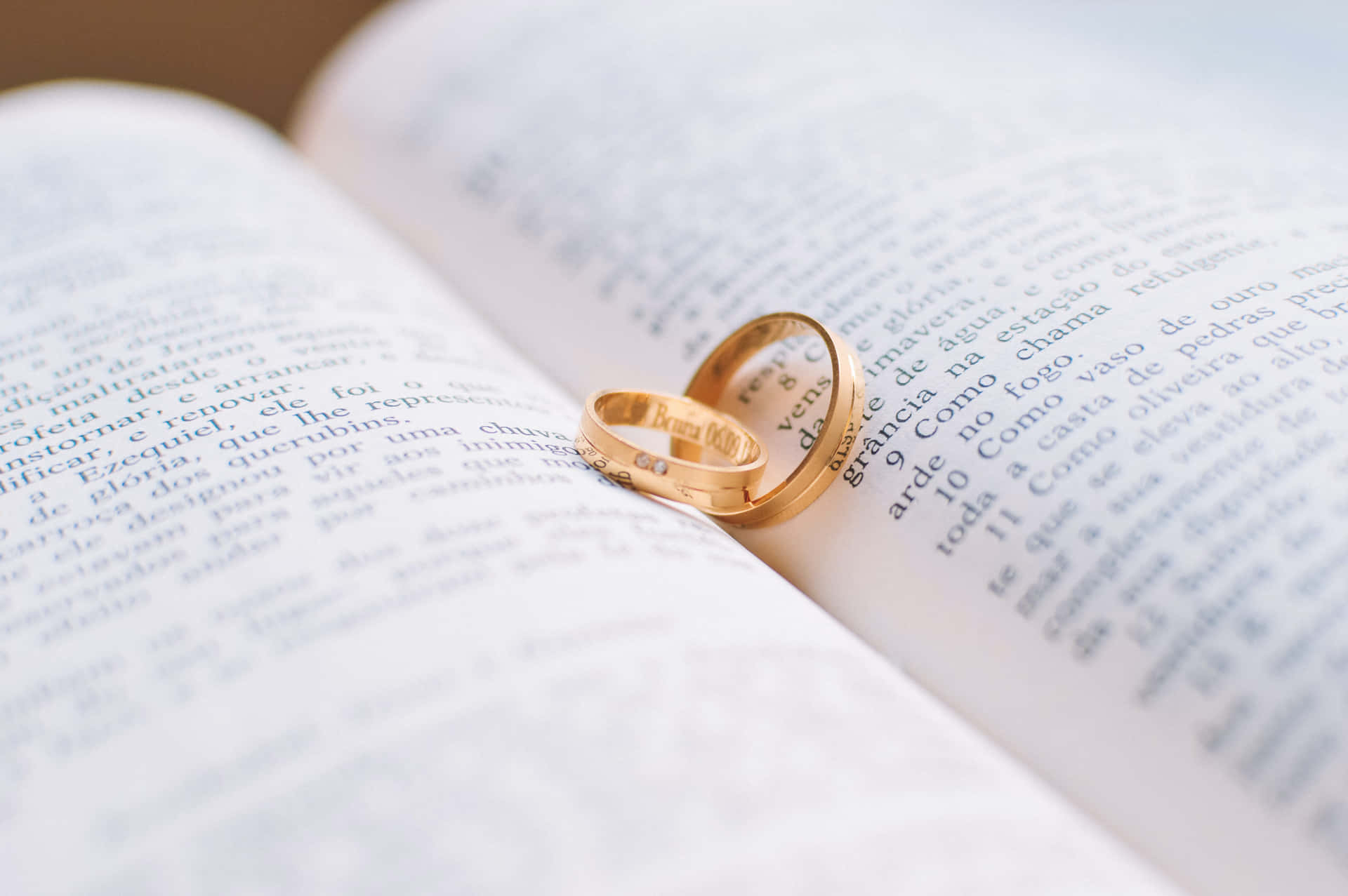 兩枚金色結婚戒指放在帶有葡萄牙語文字的打開的書頁上。 — 聖經 GIF 壁紙