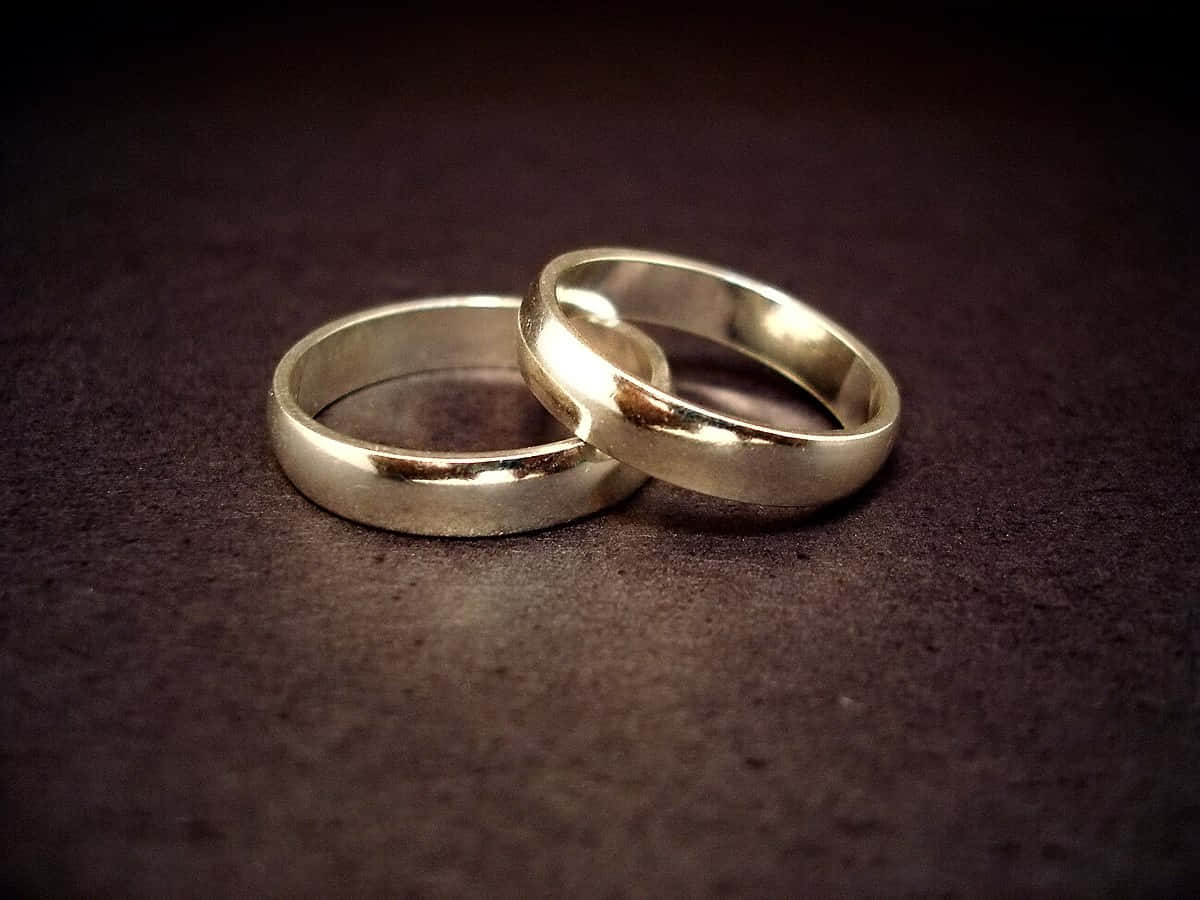 結婚戒指 桌布 壁紙 — 兩枚銀色結婚戒指在柔和燈光下交織在深色紋理表面上。