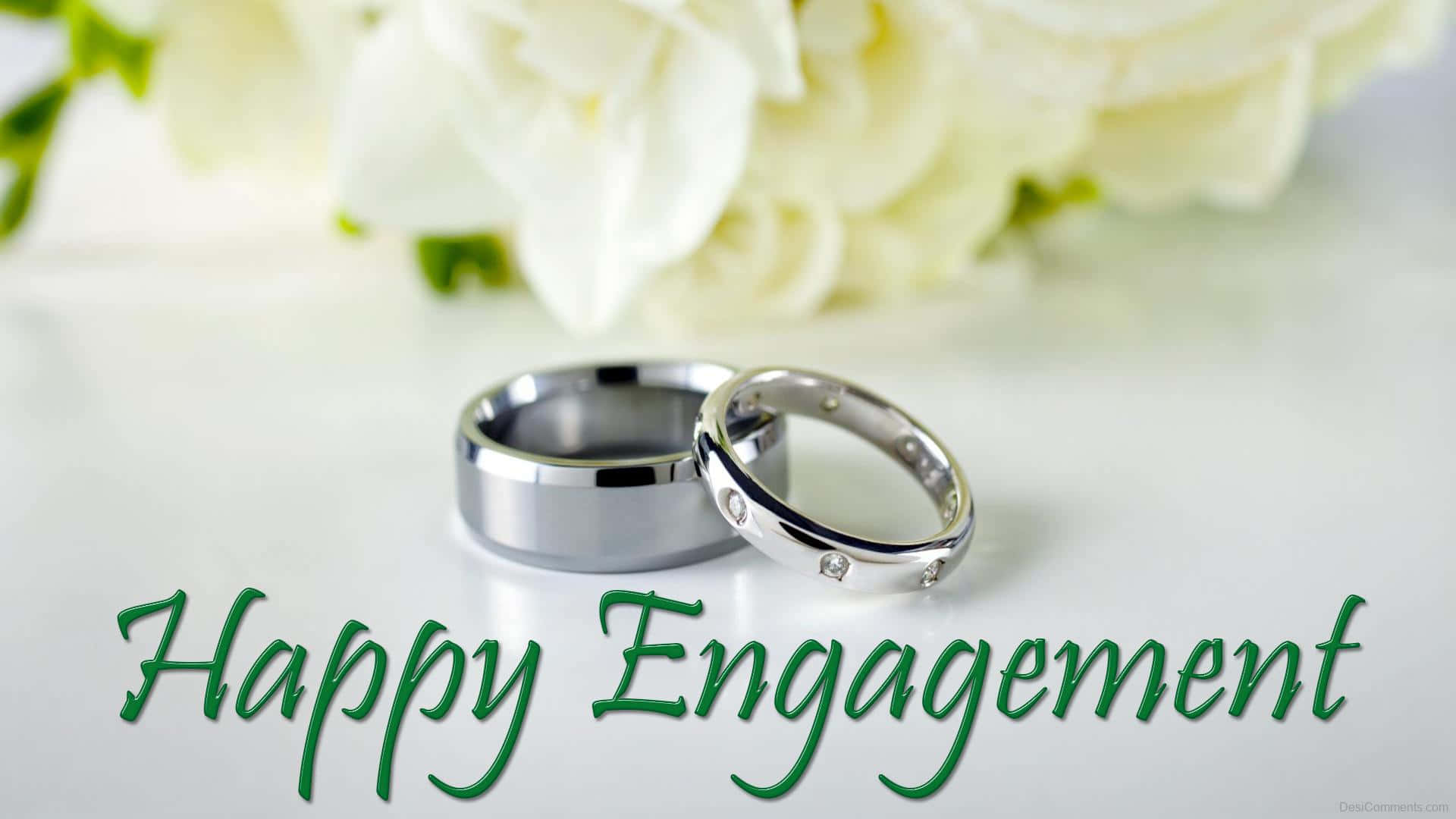 Marriage 桌布 壁紙 — 兩枚銀色訂婚戒指放在白色表面上，旁邊有柔軟的白玫瑰和綠色文字寫著 Happy Engagement。