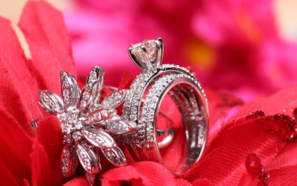 Marriage 桌布 壁紙 — 兩枚閃爍的鑽石戒指，一枚花卉造型，一枚帶狀款式，優雅地放置在鮮紅色布料上，背景是模糊的粉紅色花瓣。
