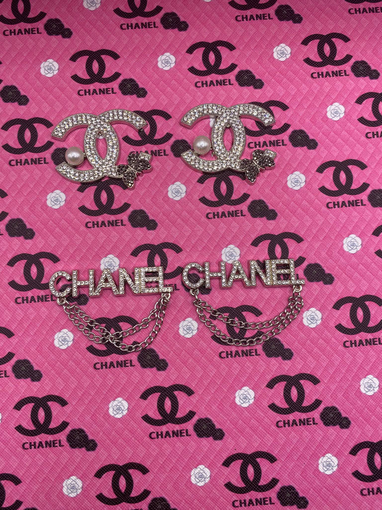 Pink Chanel Logo 桌布 壁紙 — 兩枚閃閃發光的 Chanel 胸針，飾有珍珠和鏈條，放置在帶有重複 Chanel 標誌和花卉圖案的粉紅色紋理布料上。