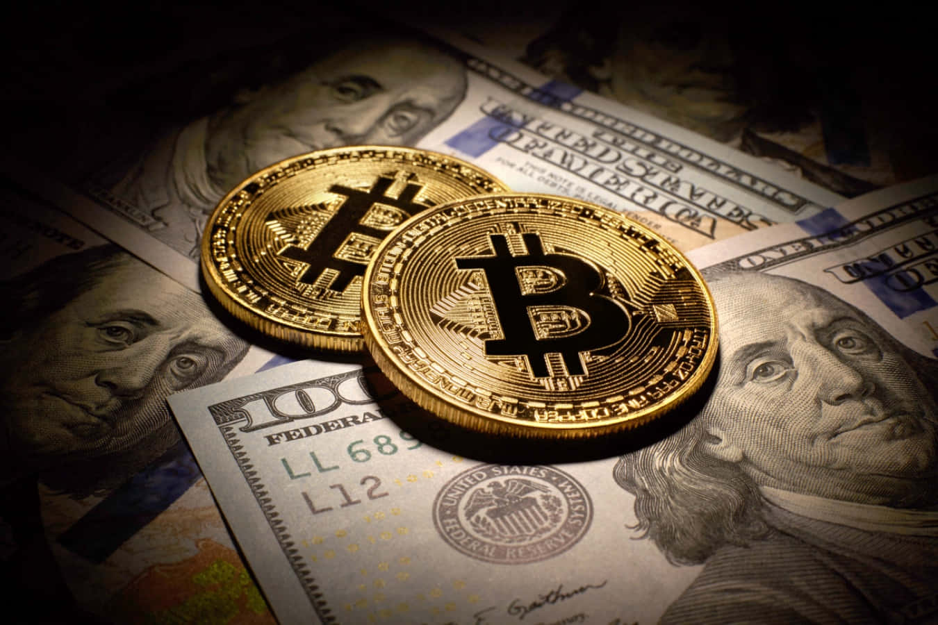 比特幣 桌布 壁紙 — 兩枚閃閃發光的金色比特幣硬幣放在一堆美元鈔票上，在戲劇性的聚光燈下。