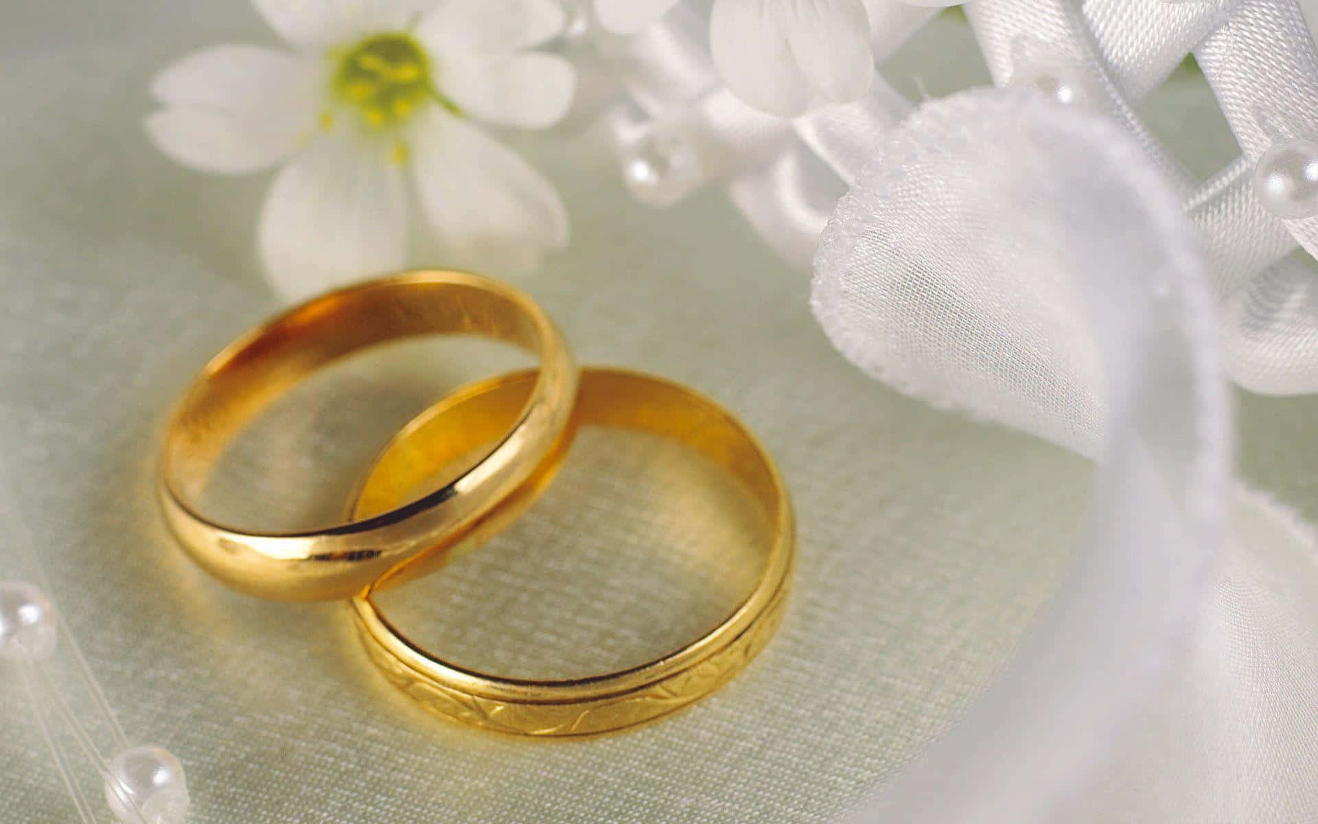 結婚戒指 桌布 壁紙 — 兩枚閃閃發光的金色結婚戒指，旁邊擺放著精緻的白色花朵和透明絲帶，置於柔軟的紋理表面上。