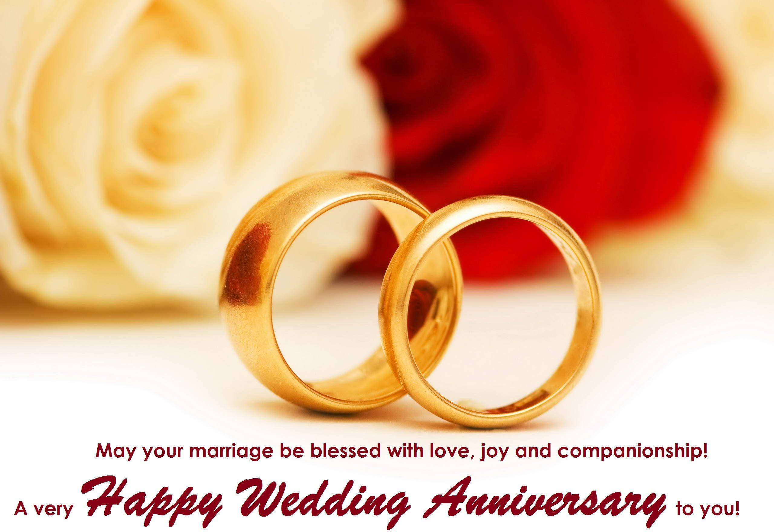 Happy Anniversary 桌布 壁紙 — 兩枚閃閃發光的金色結婚戒指交織在一起，背景是柔和的紅玫瑰和奶油色玫瑰，並帶有慶祝週年紀念的文字。