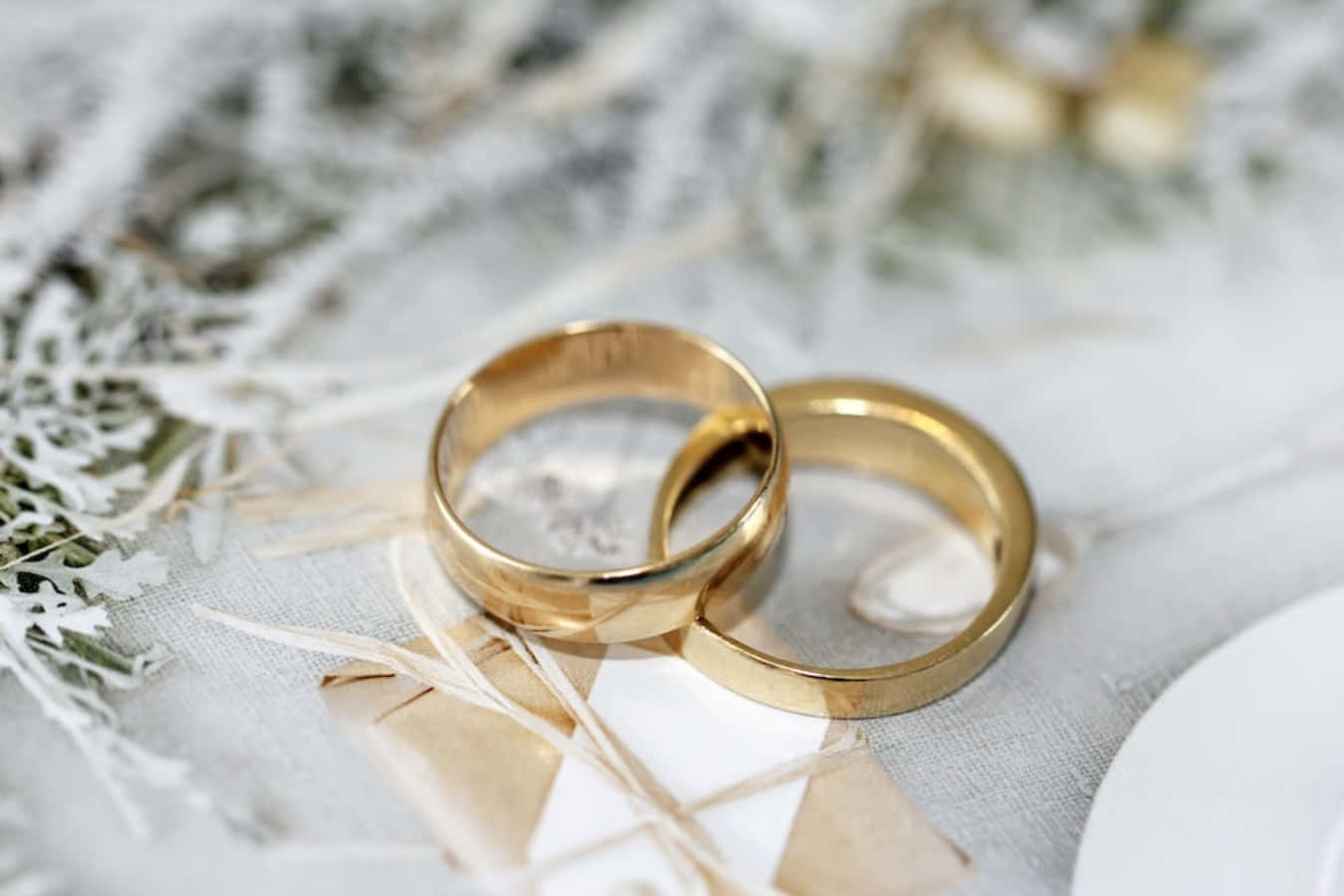 結婚戒指 桌布 壁紙 — 兩枚閃閃發光的金色結婚戒指放在柔軟的白色布料上，旁邊有乾燥的花卉裝飾。