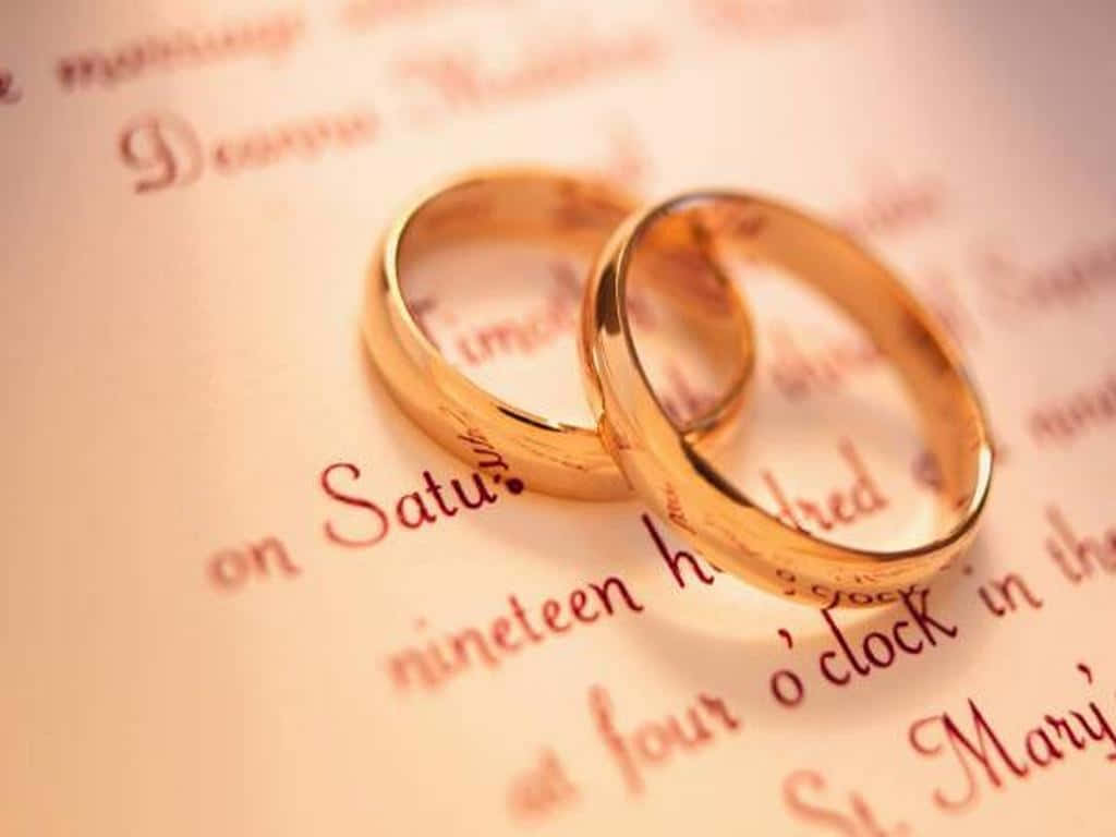結婚戒指 桌布 壁紙 — 兩枚閃閃發光的金色結婚戒指親密地放在一張帶有優雅草書字體的復古婚禮邀請函上。