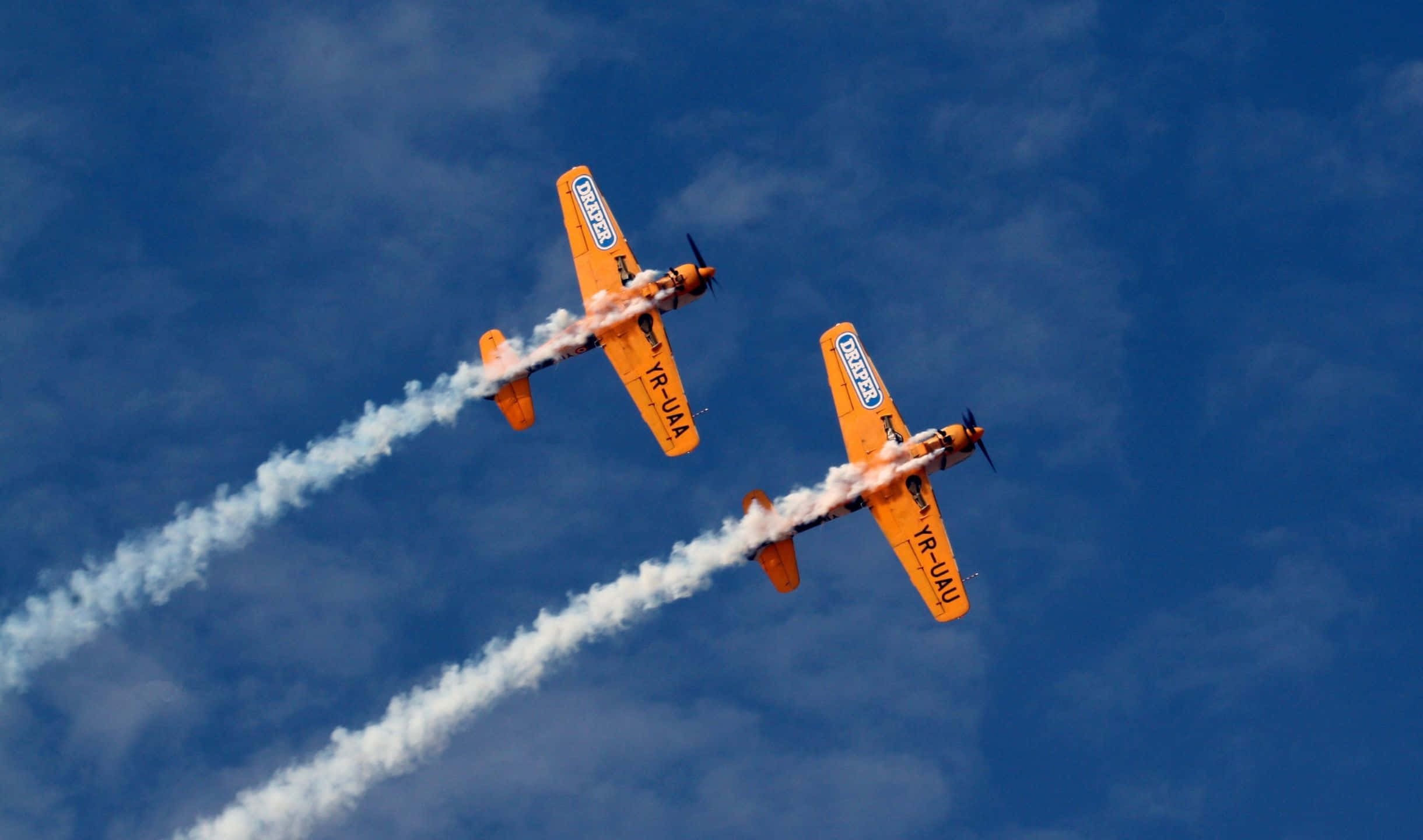 2440x1440 Small Planes 桌布 壁紙 — 兩架亮橙色特技飛機對角線飛過深藍色天空，身後留下濃厚白色煙跡。