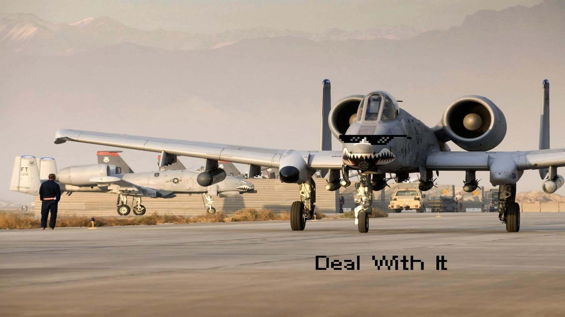 A 10 Warthog 桌布 壁紙 — 兩架帶有鯊魚嘴貼花的軍用噴射機停在塵土飛揚的機場上，背景朦朧中有積雪的山峰。