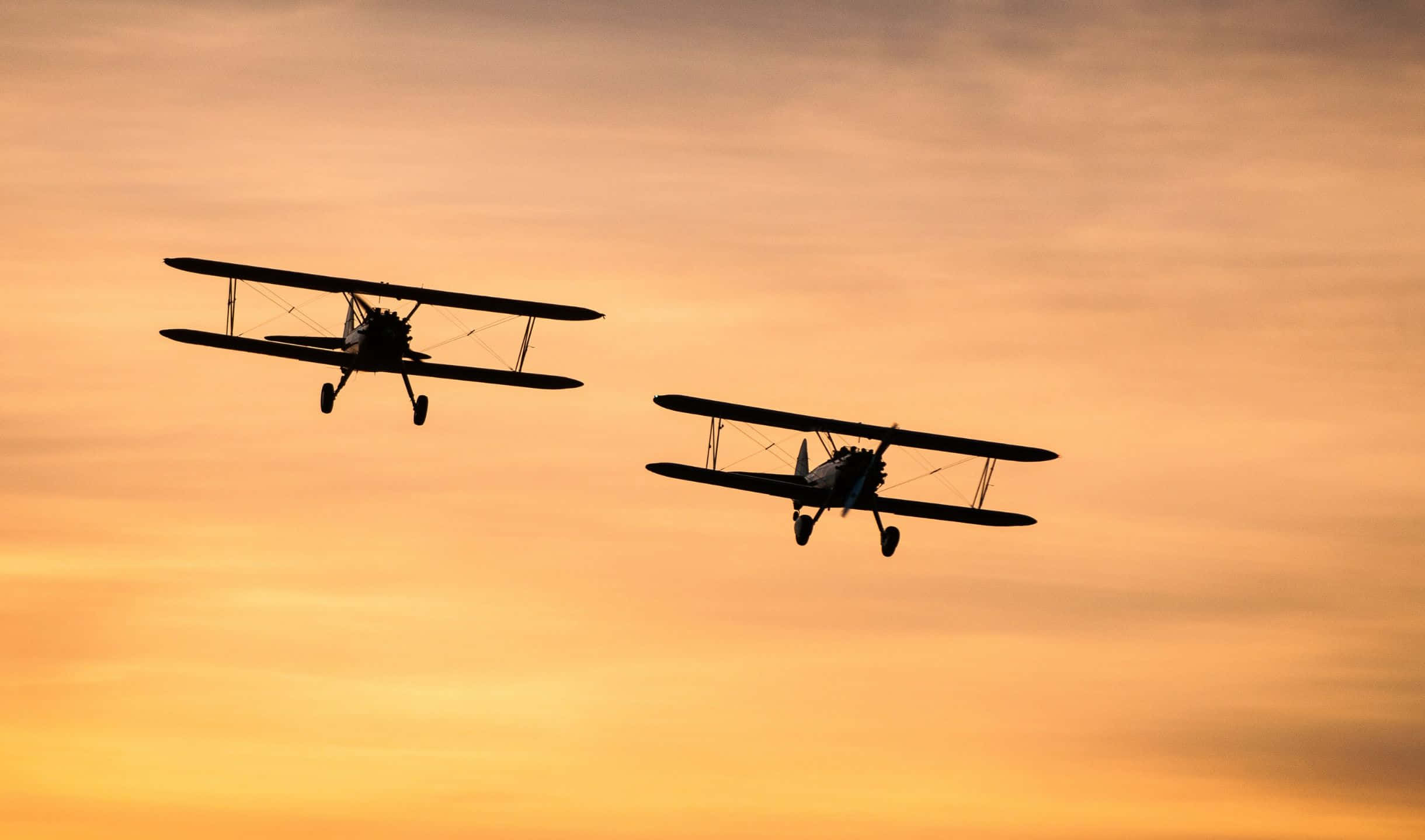 2440x1440 Small Planes 桌布 壁紙 — 兩架復古雙翼機在溫暖的金色日落天空背景下以剪影翱翔。