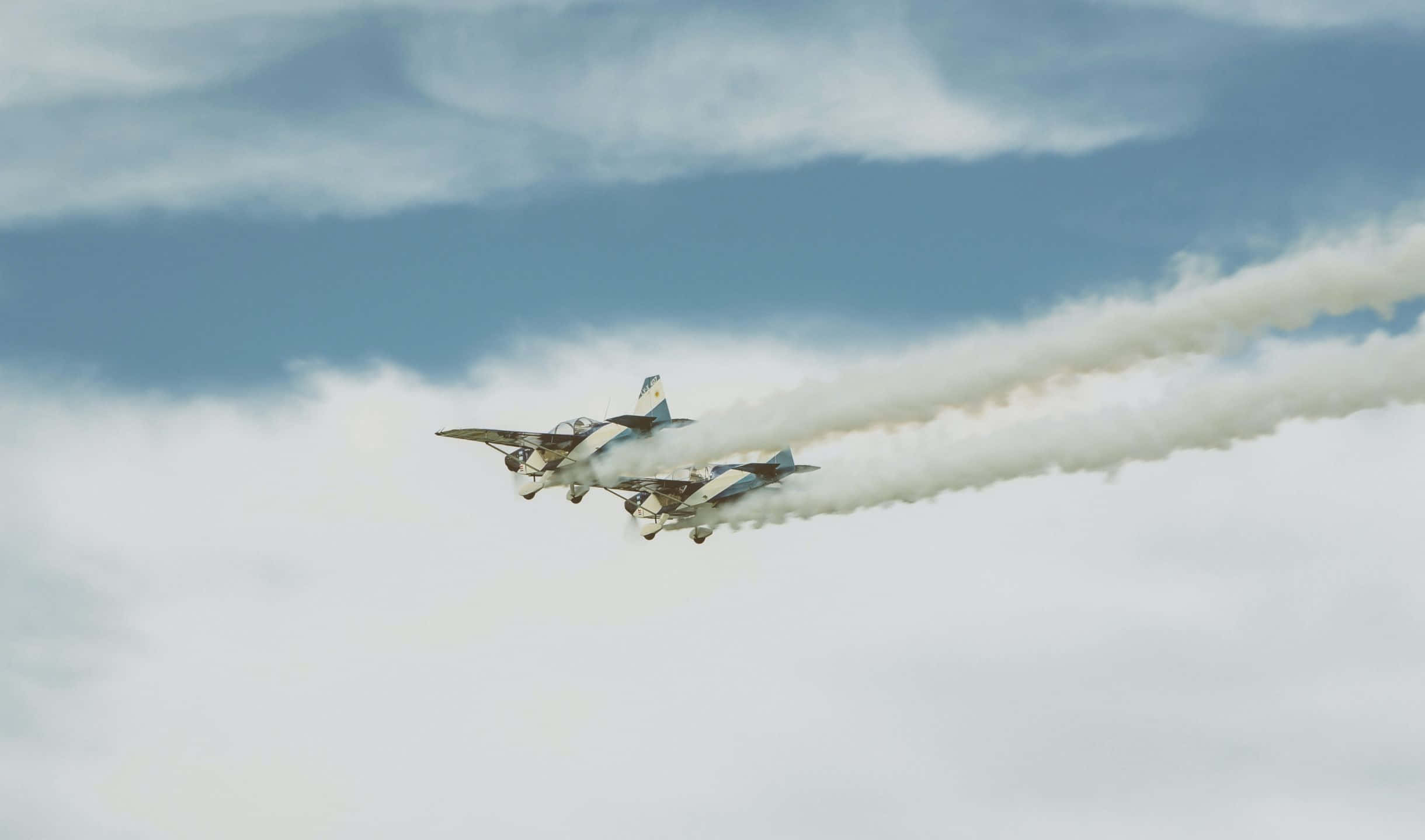 2440x1440 Small Planes 桌布 壁紙 — 兩架復古雙翼機編隊飛過多雲的天空，留下濃厚的白色煙霧痕跡。