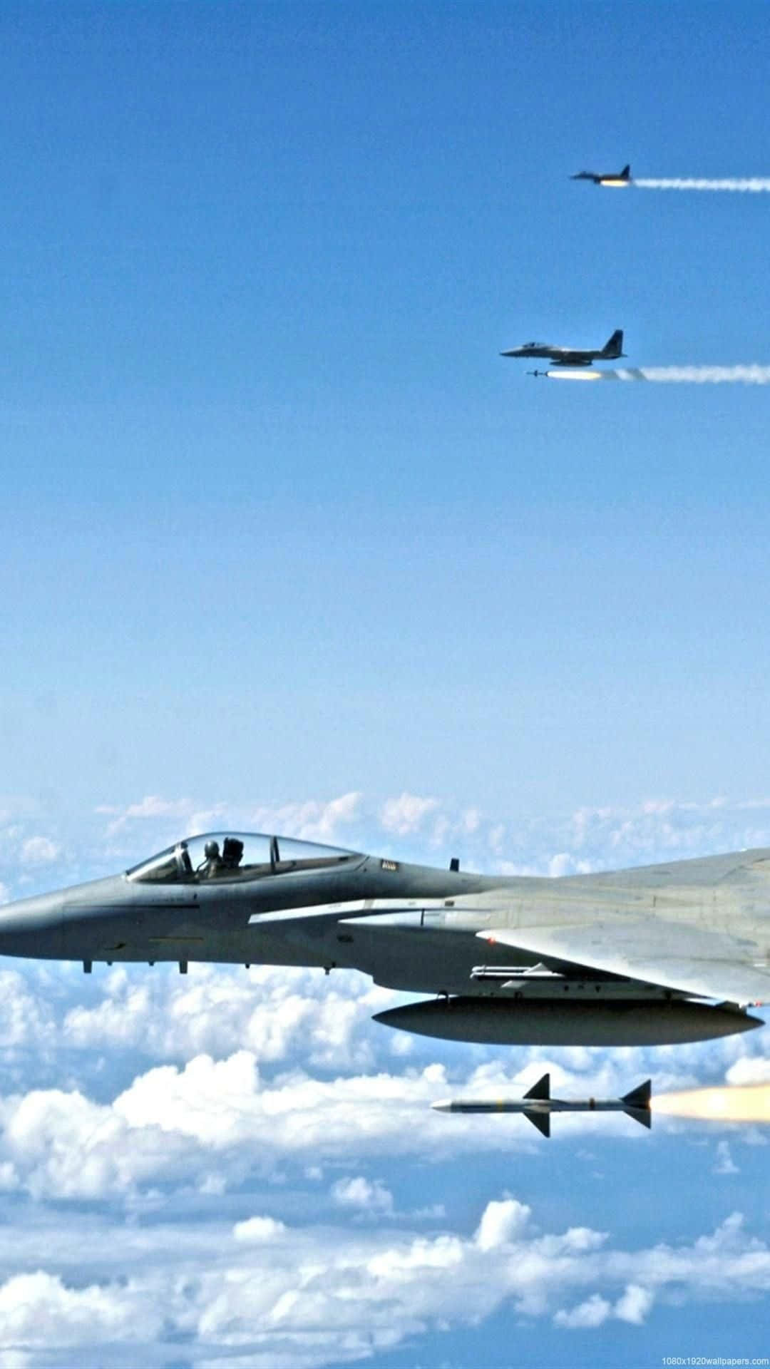 Android Small Planes 桌布 壁紙 — 兩架戰鬥機在清澈的藍天中翱翔，飛越蓬鬆的白雲，其中一架發射飛彈，留下煙霧痕跡。