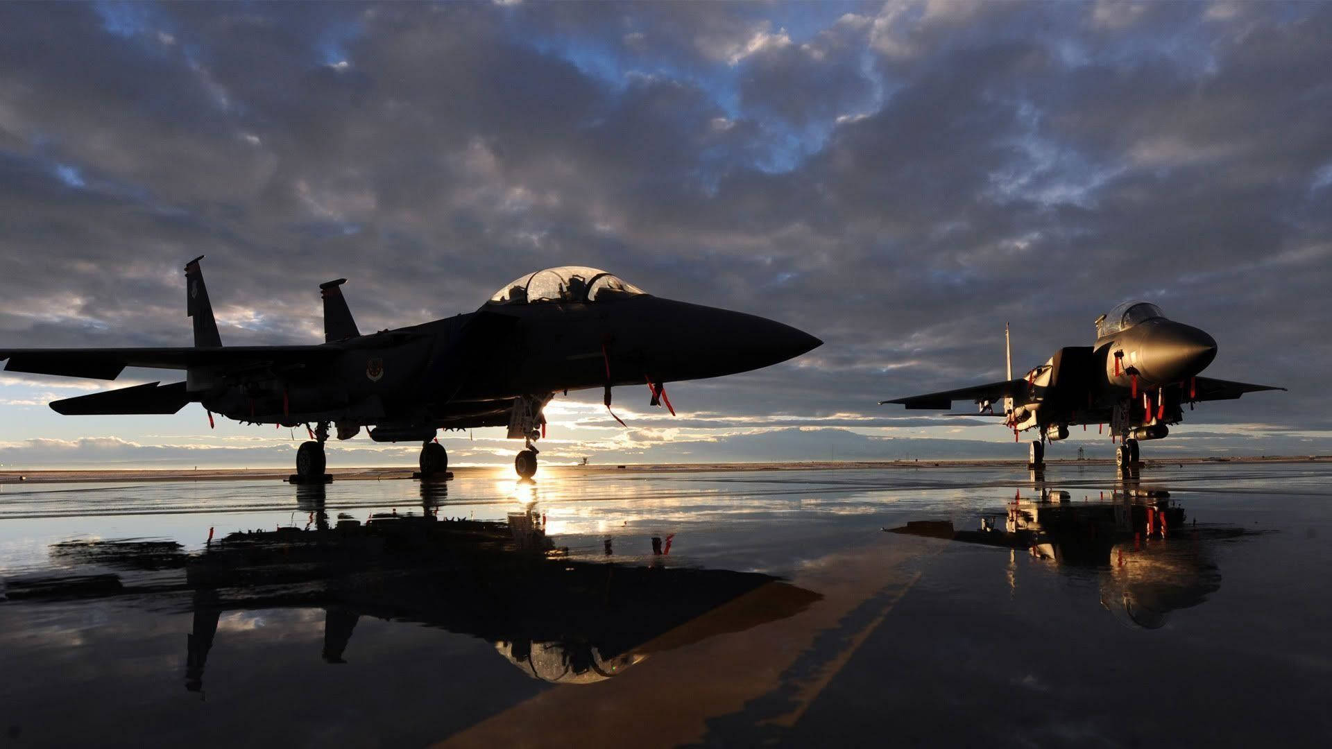 飛機 桌布 壁紙 — 兩架戰鬥機在濕滑的停機坪上形成剪影，夕陽在它們身後落下，投射出戲劇性的倒影。