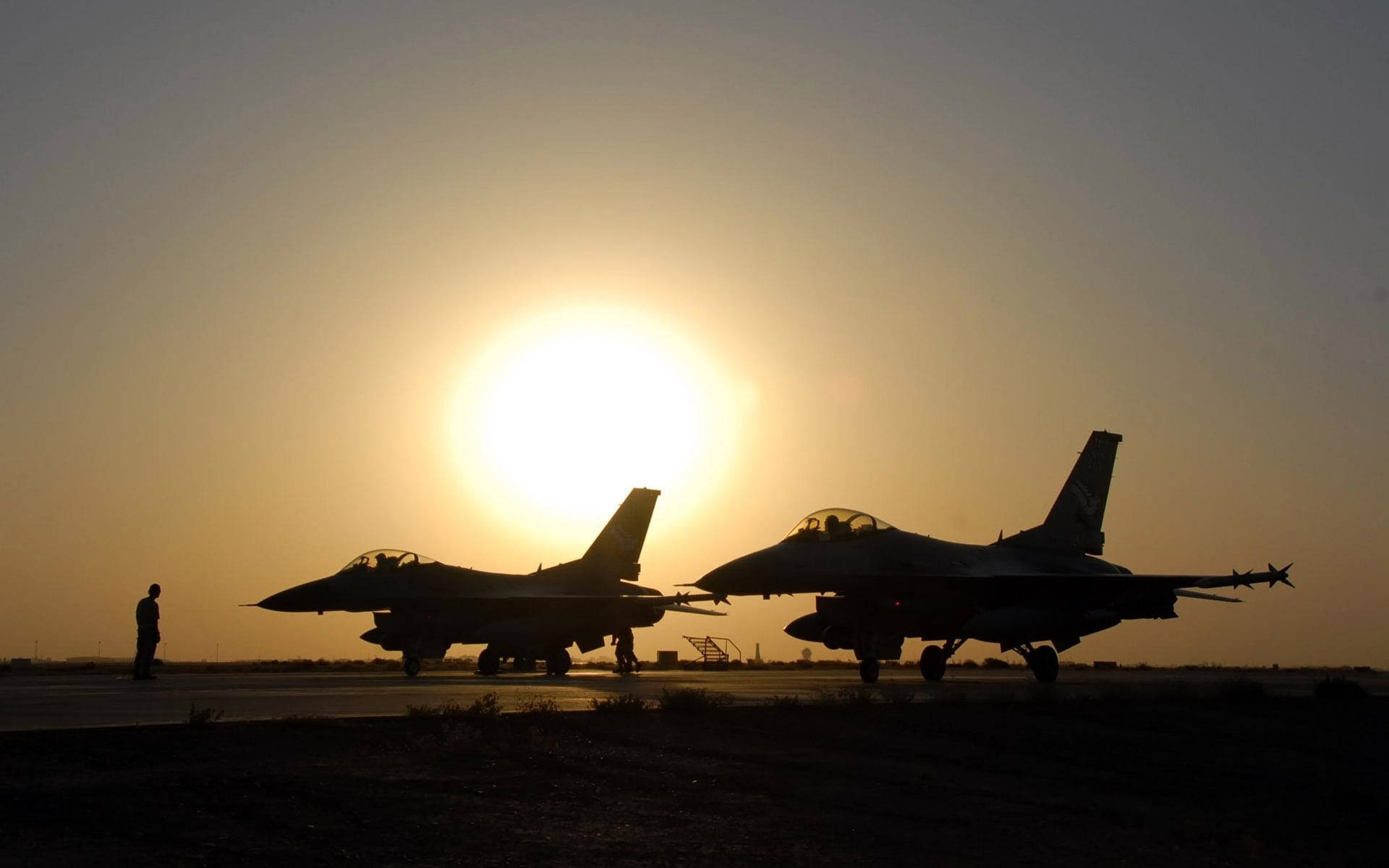 軍事動圖 桌布 壁紙 — 兩架戰鬥機在跑道上以剪影站立，明亮的太陽在它們身後落下，投下長長的影子。