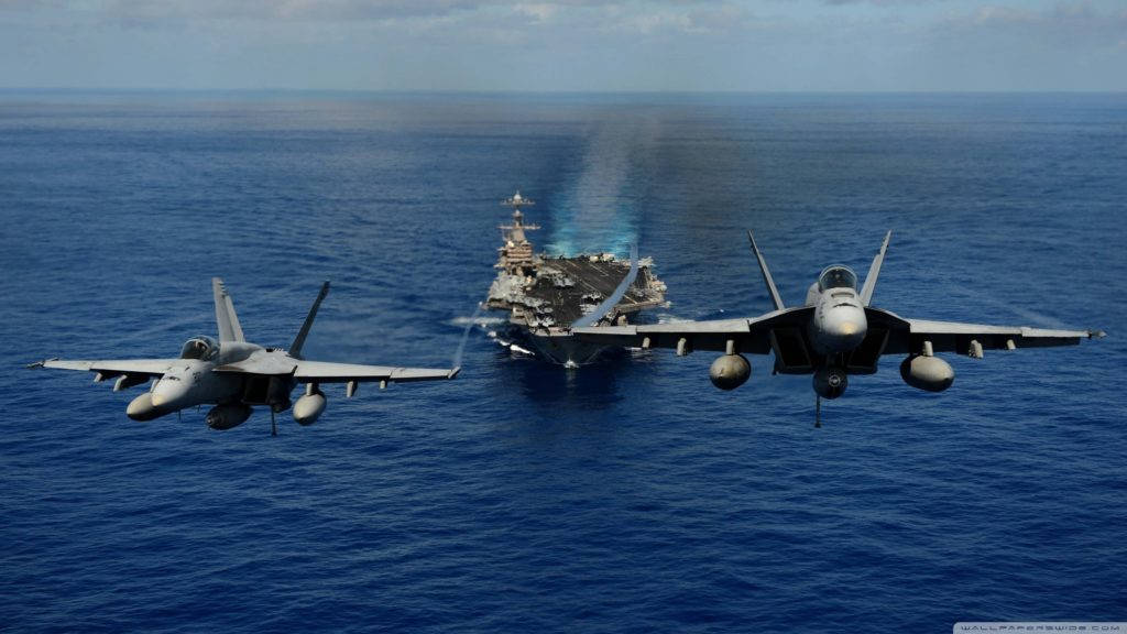 u s navy 桌布 壁紙 — 兩架戰鬥機編隊飛行在廣闊的藍色海洋上空，遠處有一艘航空母艦拖著尾跡。