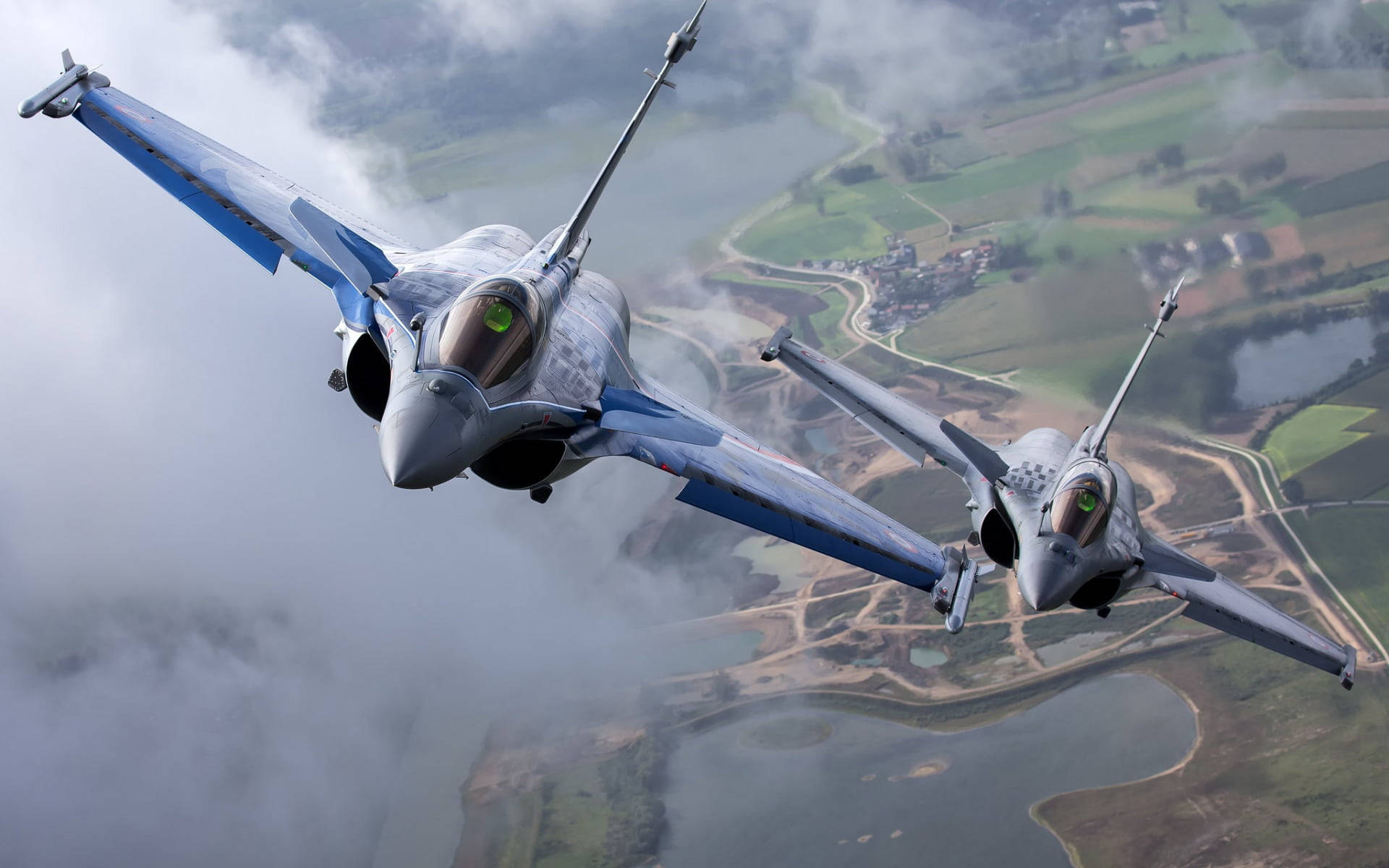 軍事動圖 桌布 壁紙 — 兩架流線型戰鬥機在雲層中飛馳，下方是綠色田野和蜿蜒河流組成的拼布般景觀。