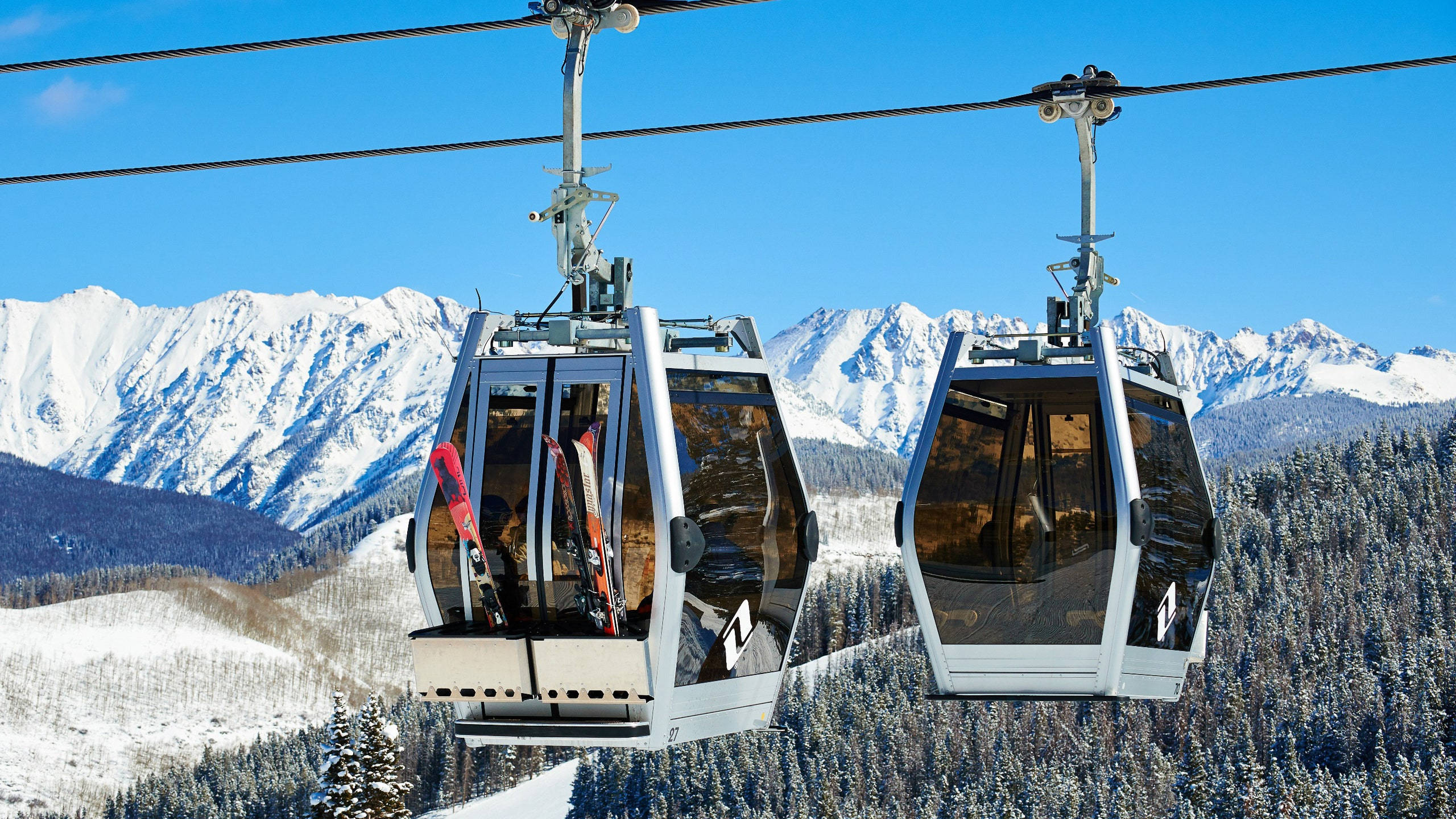 Colorado 桌布 壁紙 — 兩架滑雪纜車在晴朗藍天下，於積雪山脈和常綠森林上方滑行。