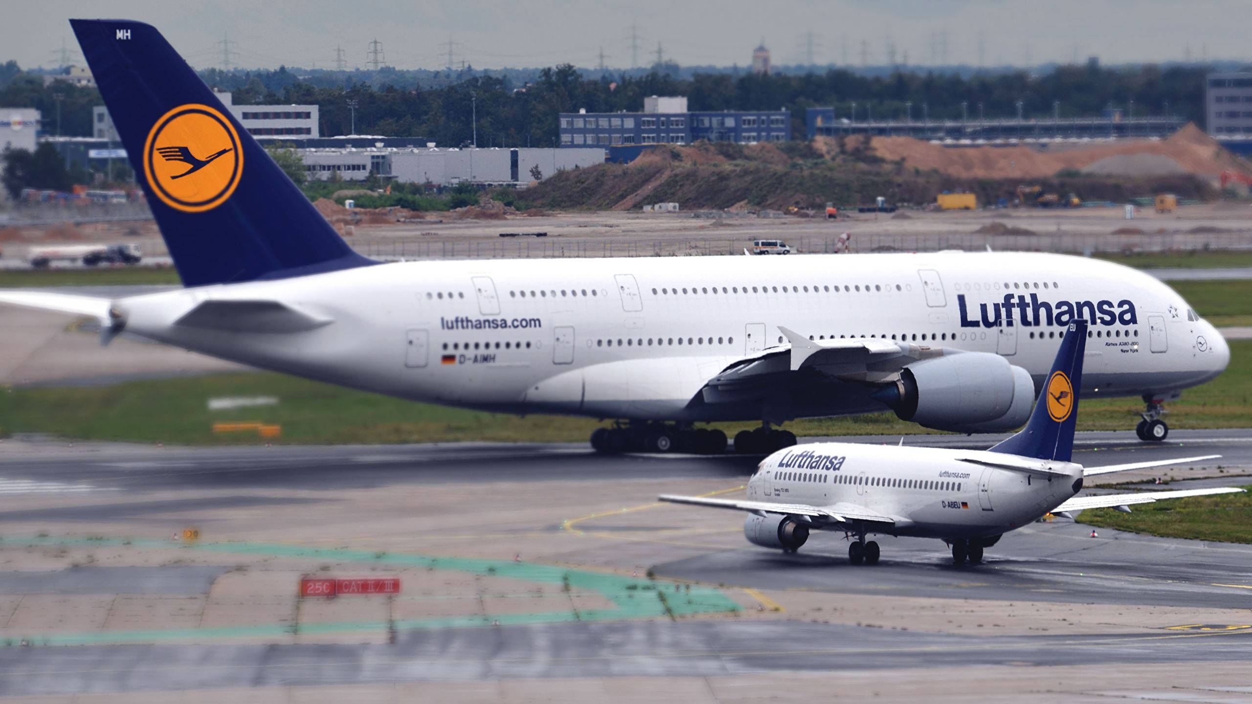 飛機 桌布 壁紙 — 兩架漢莎航空飛機在機場停機坪，一大一小，背景有跑道和建築物。