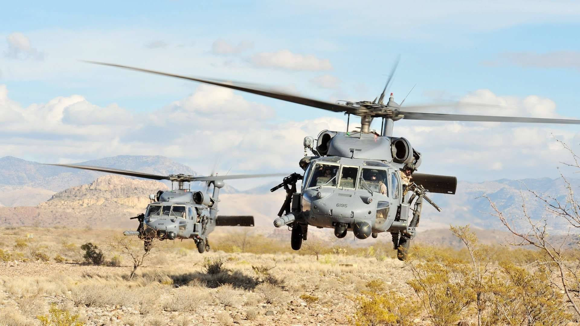 Black Hawk Helicopter 桌布 壁紙 — 兩架灰色軍用直升機在乾燥的沙漠景觀上空低空盤旋，遠方有山脈，天空局部多雲。