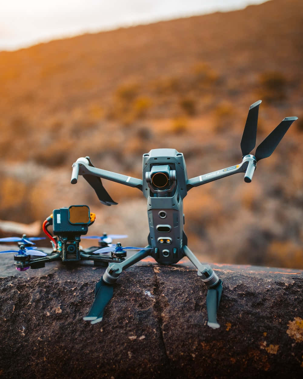 drone 桌布 壁紙 — 兩架無人機停在一塊崎嶇的岩石上，溫暖的金色光芒沐浴著背景中的沙漠山坡。