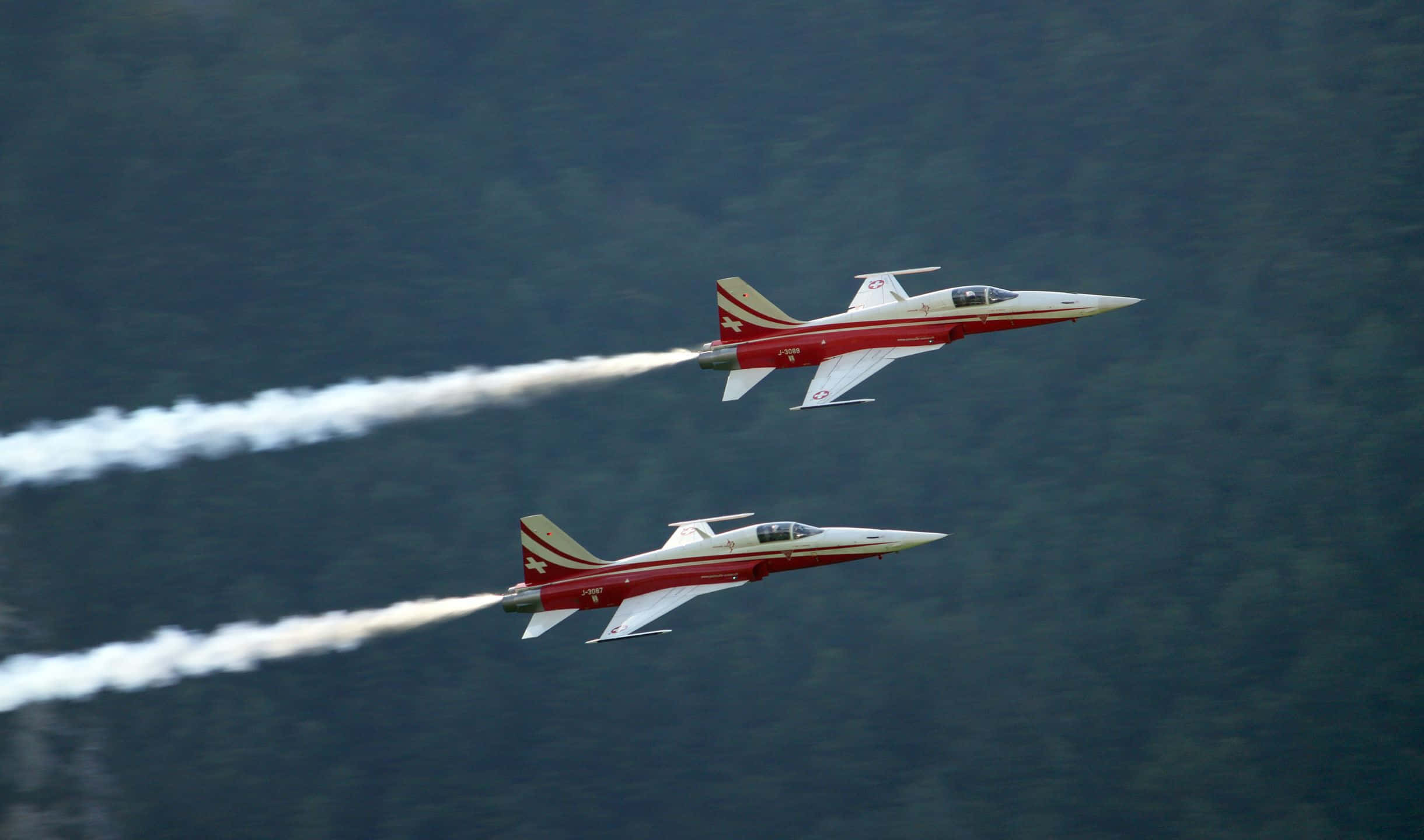 2440x1440 Small Planes 桌布 壁紙 — 兩架紅白相間的戰鬥機以緊密編隊飛越茂密的綠色森林，留下白色煙霧痕跡。