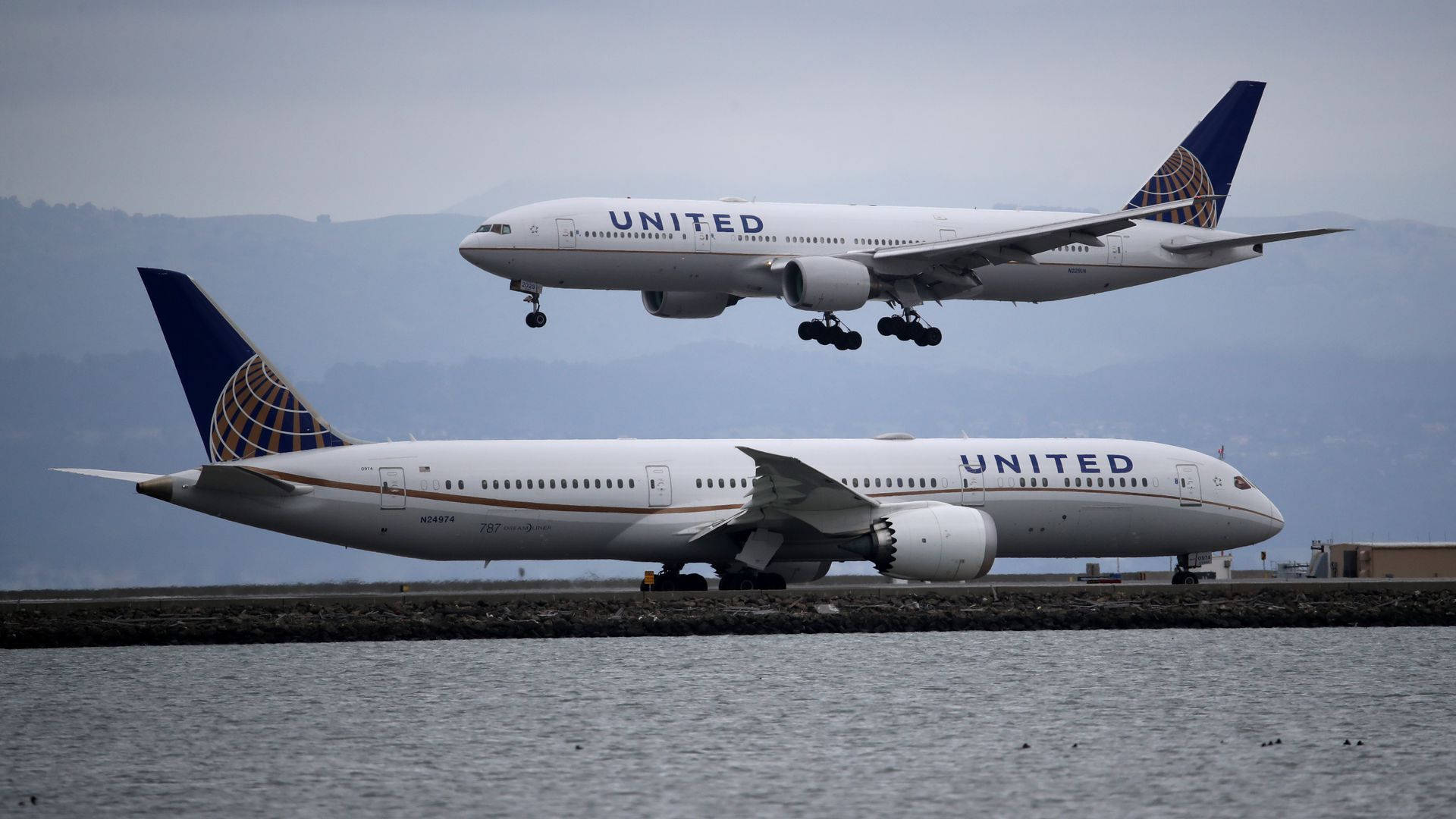 飛機 桌布 壁紙 — 兩架聯合航空噴射機，一架在空中放下起落架，另一架在水邊滑行，背後有朦朧的山丘。