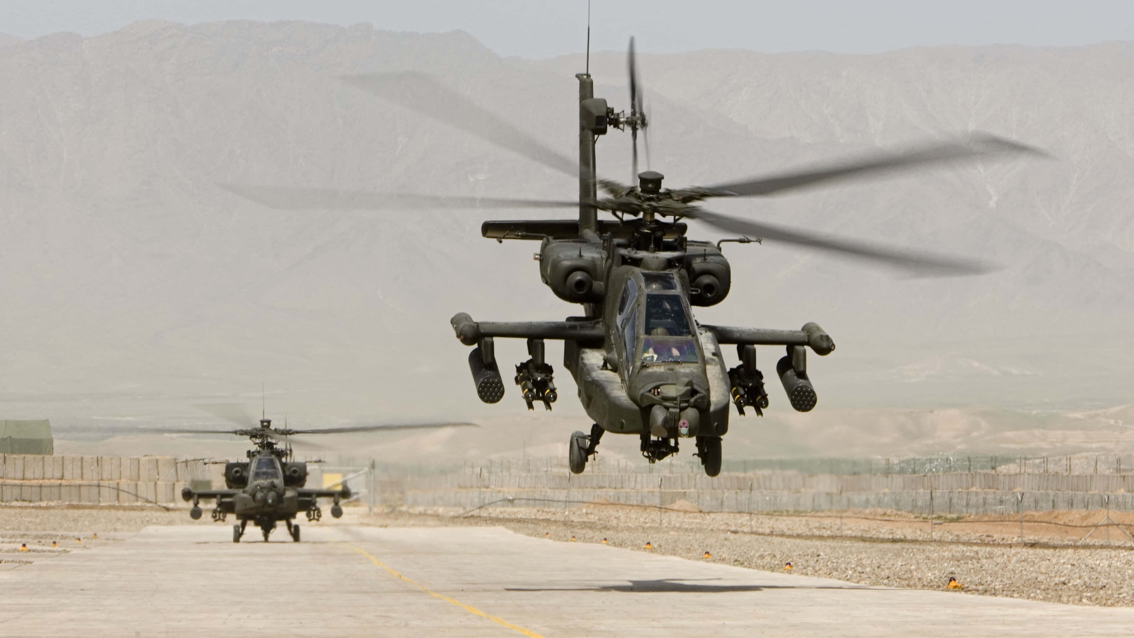 4K Helicopters 桌布 壁紙 — 兩架軍用攻擊直升機在塵土飛揚的機場上空盤旋並滑行，背景是朦朧的崎嶇山脈。